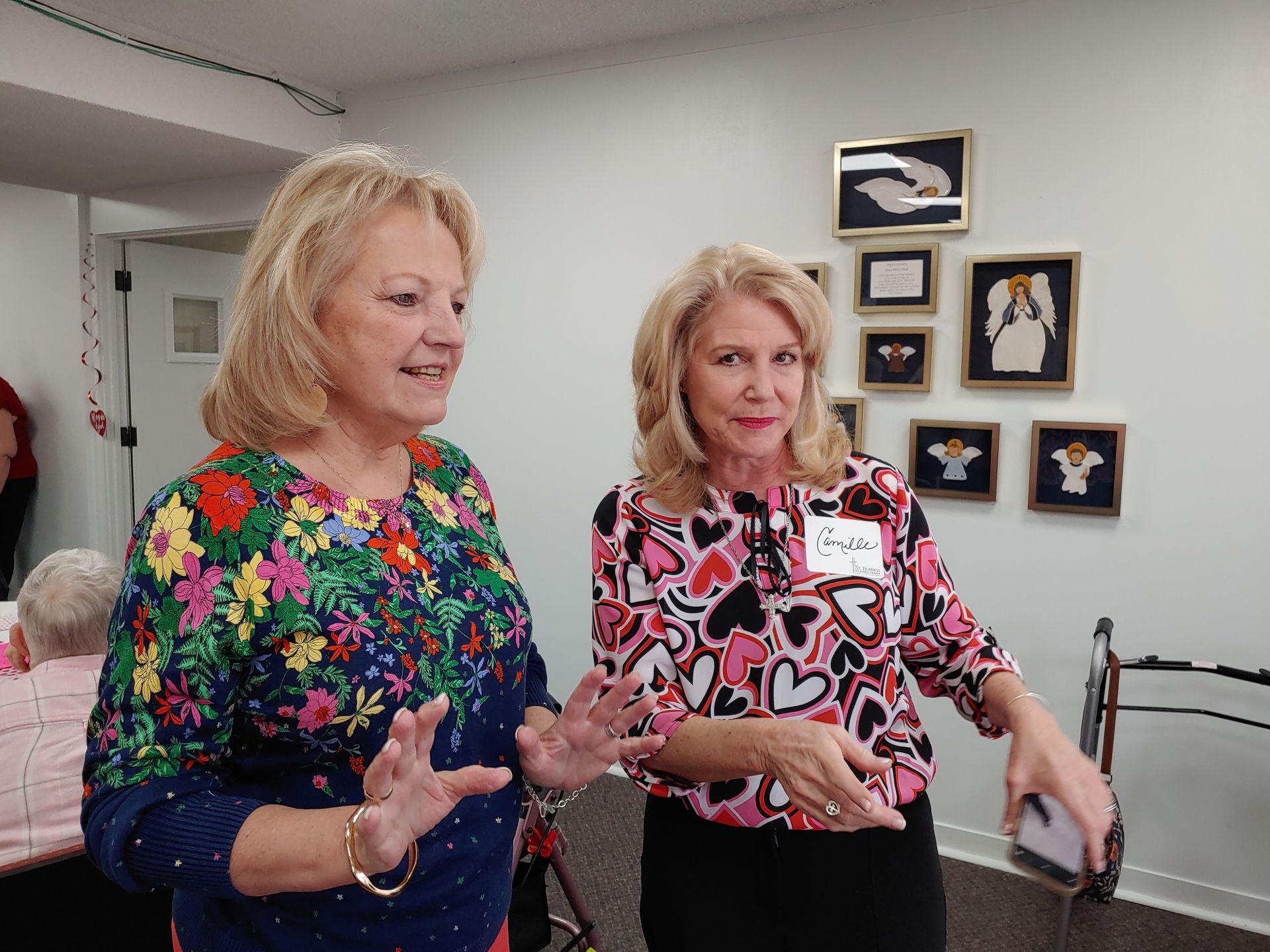 Two women talking, one in floral top, the other in heart print, near framed art on a white wall.