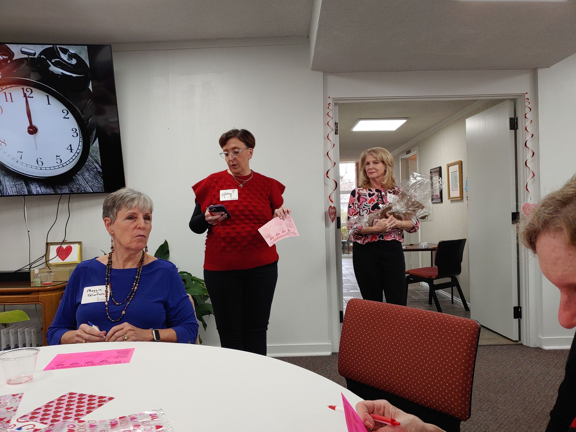 People at a gathering, one woman speaks, another holds a basket, papers on a table.