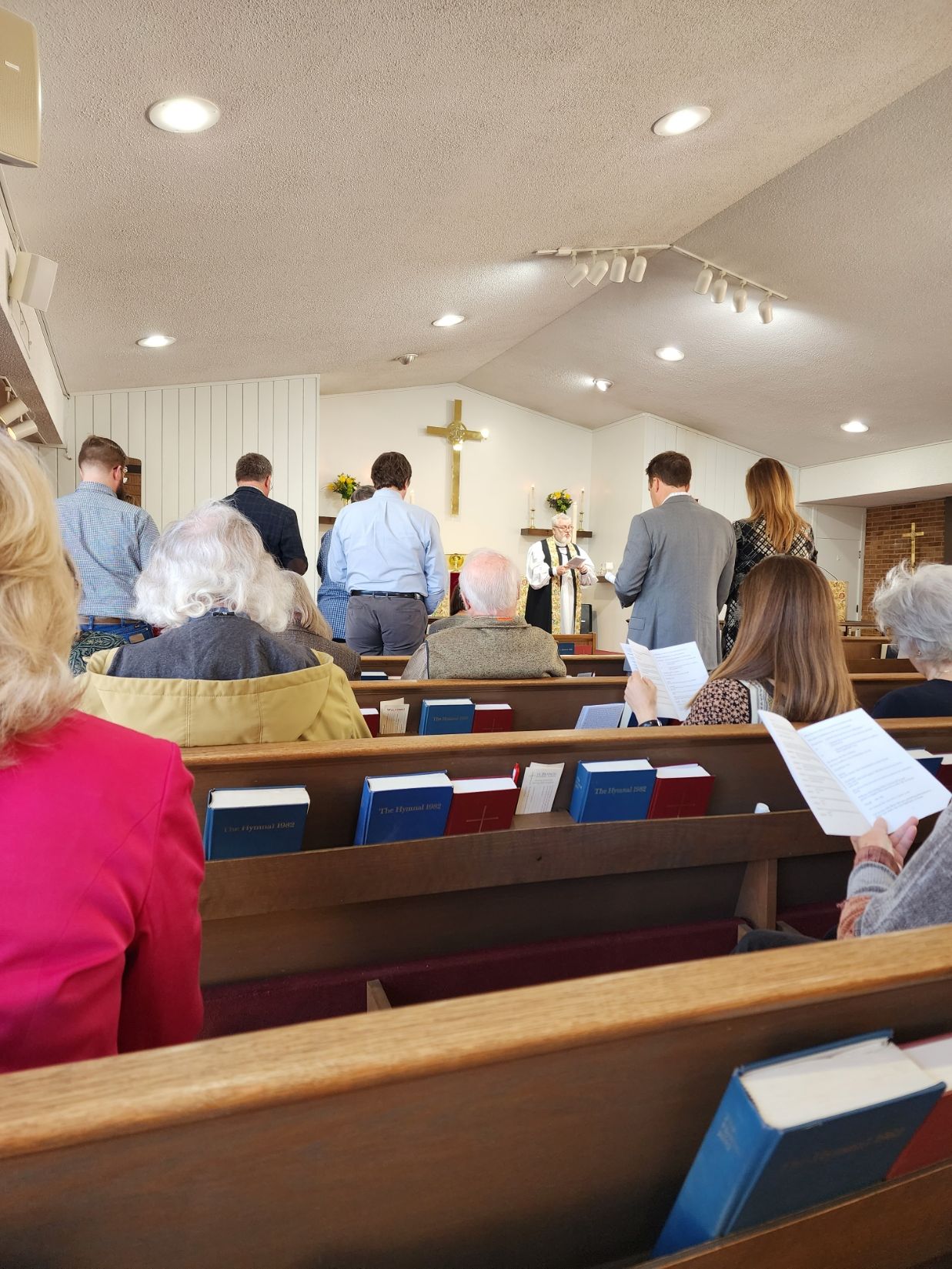 People standing in a church service; pews in foreground; cross above altar.