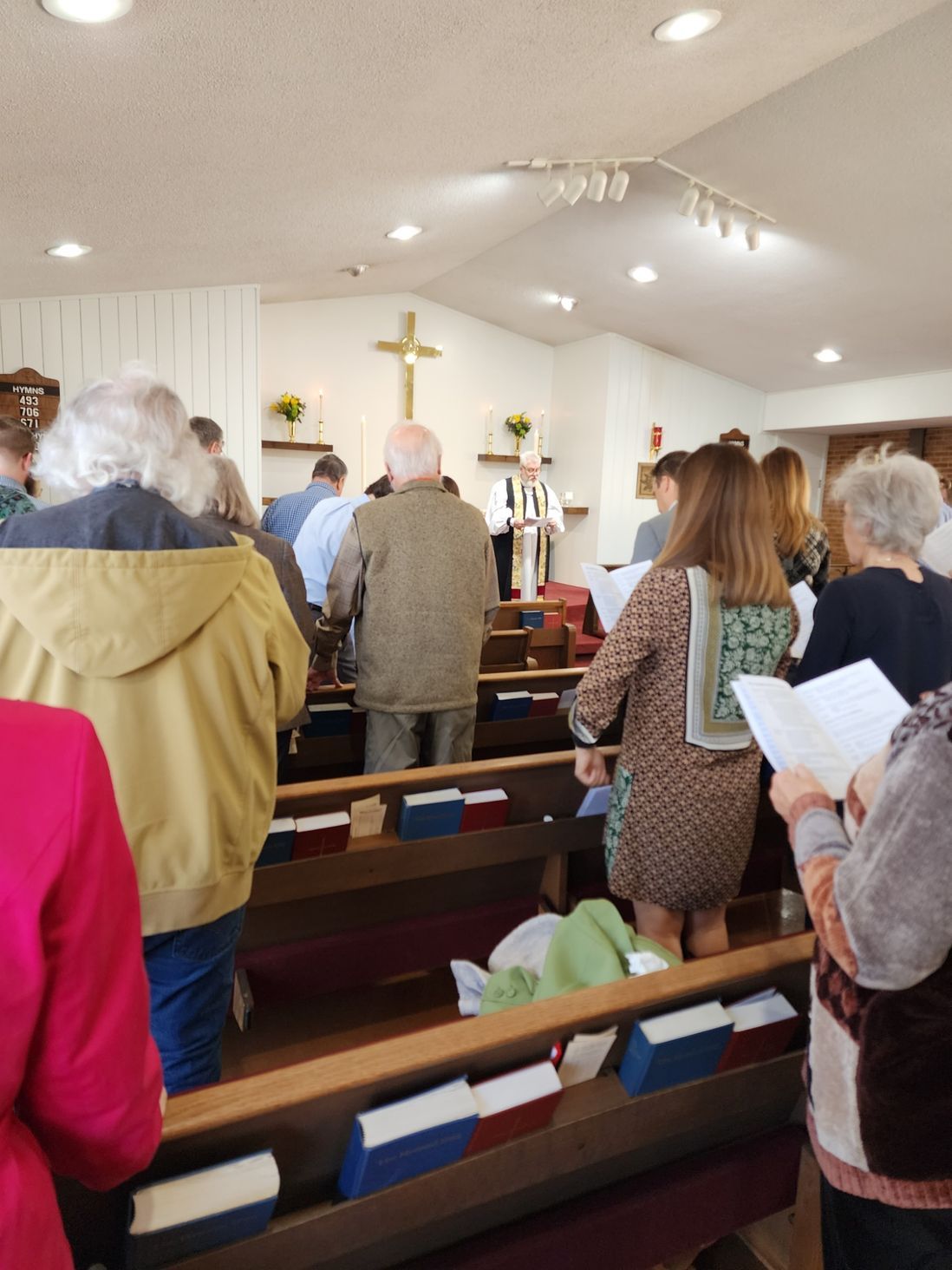 People standing and sitting in a church, facing a cross, some holding songbooks.