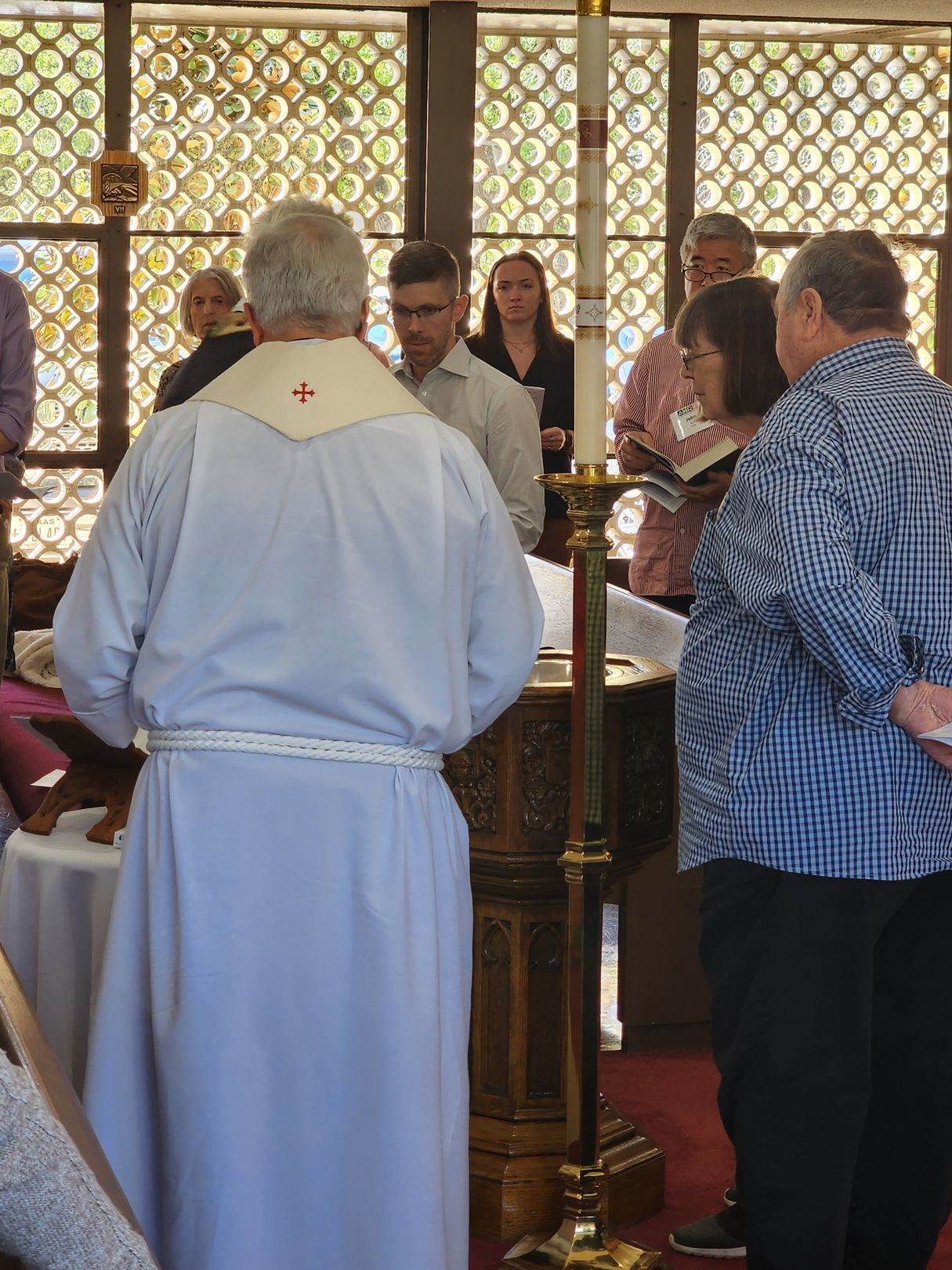 A baptism ceremony. A person in white robes stands at a baptismal font. Others are standing and looking on.