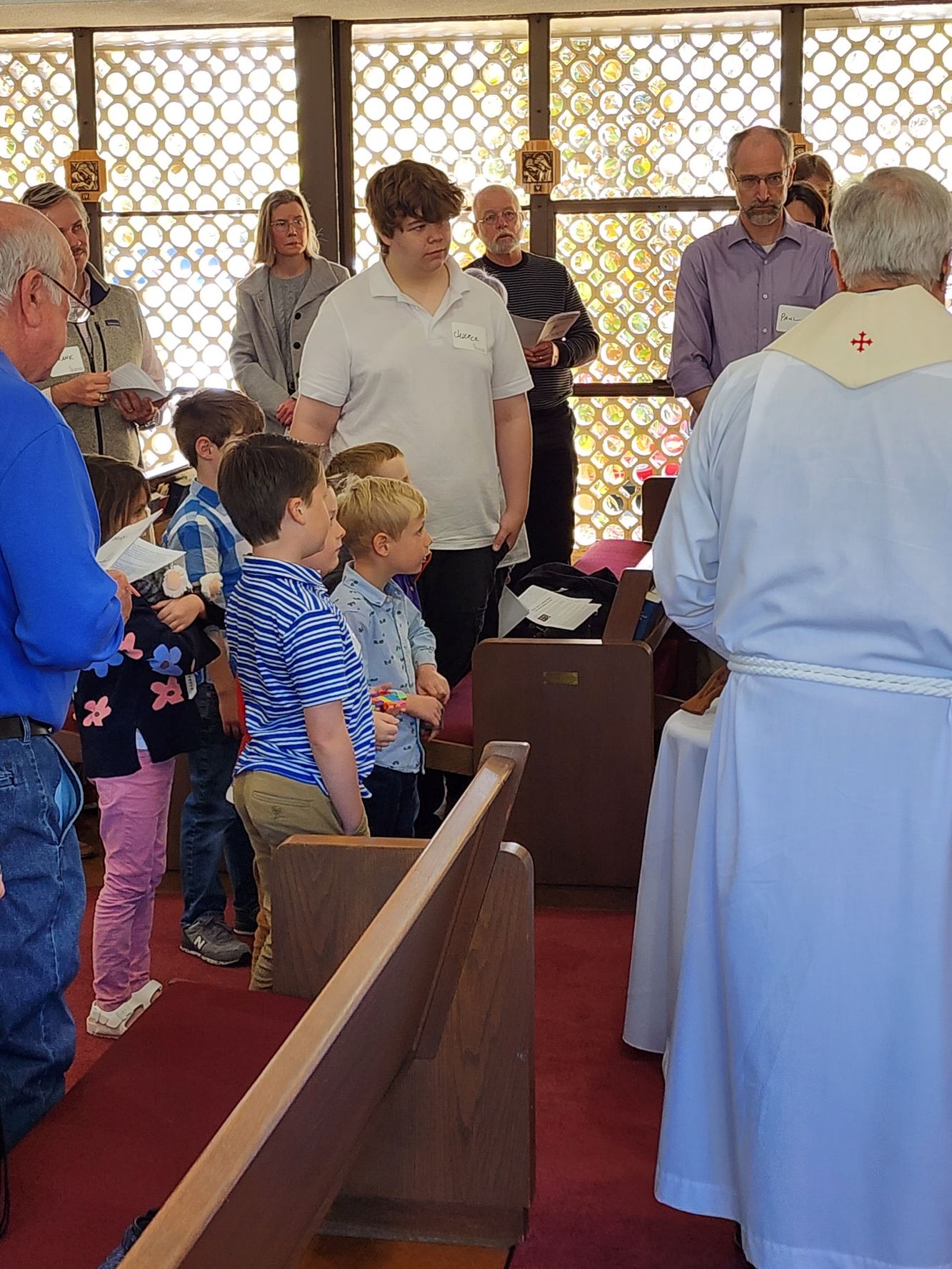 People in a church gather near a priest in a white robe. Children stand close, while others look on.
