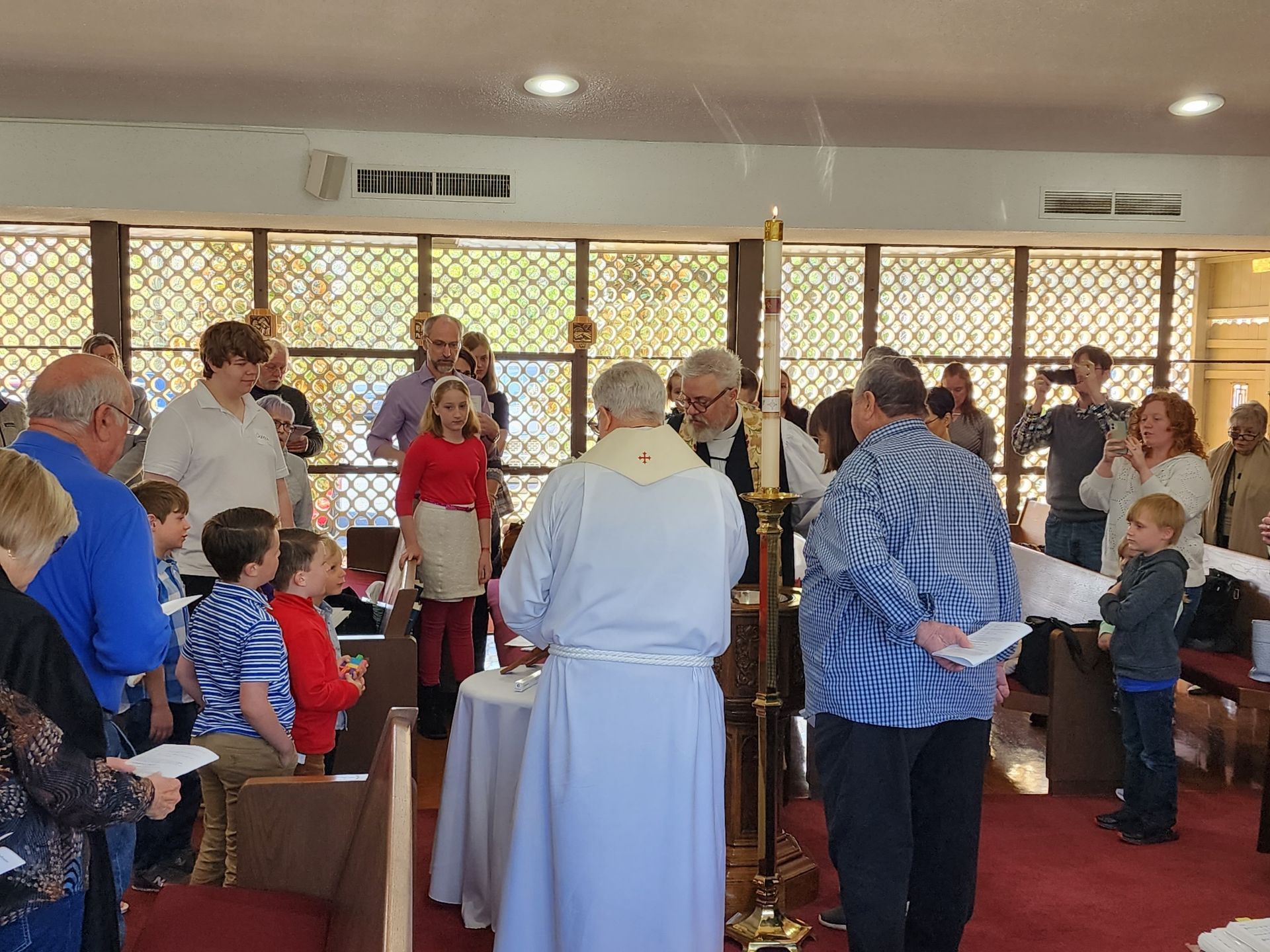 People gathered in church for a ceremony. A priest stands at a table, people hold papers, and a tall candle is lit.