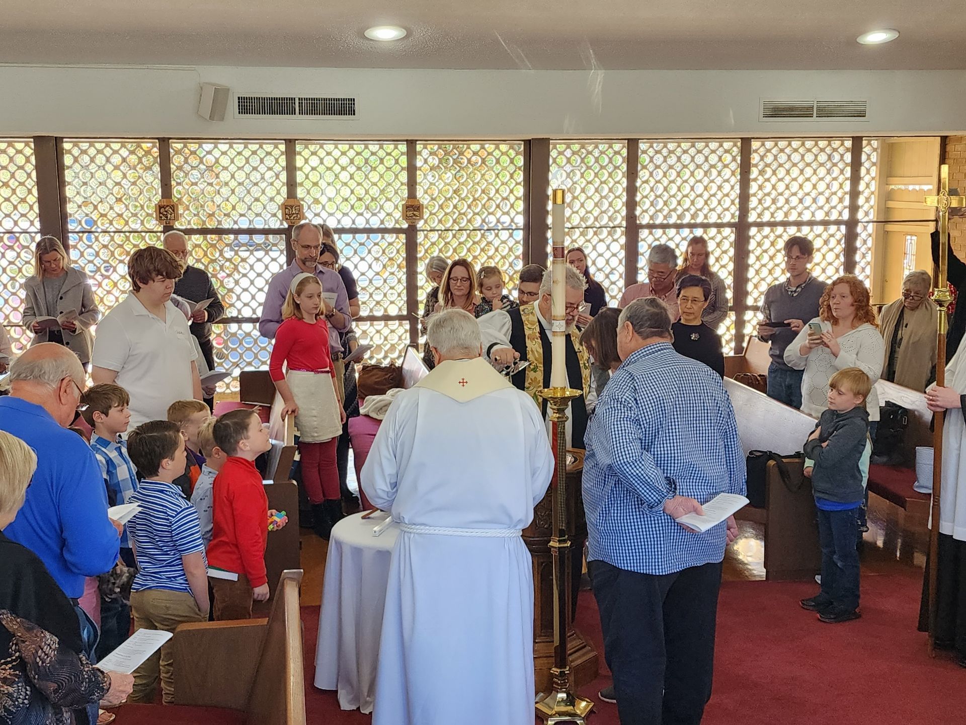 People in a church service. A priest in white vestments is at the altar, surrounded by people of different ages.