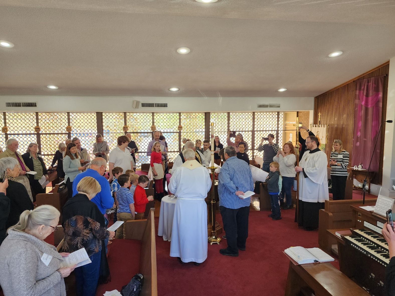 People gather in a church. Sunlight streams through windows. Individuals read from booklets, with a person in white officiating.