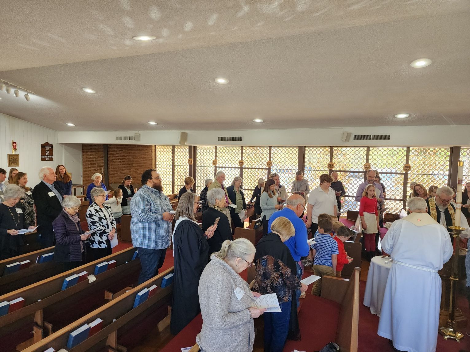 People at a church service; some stand, some seated in pews. Priest at an altar, sunlight through windows.