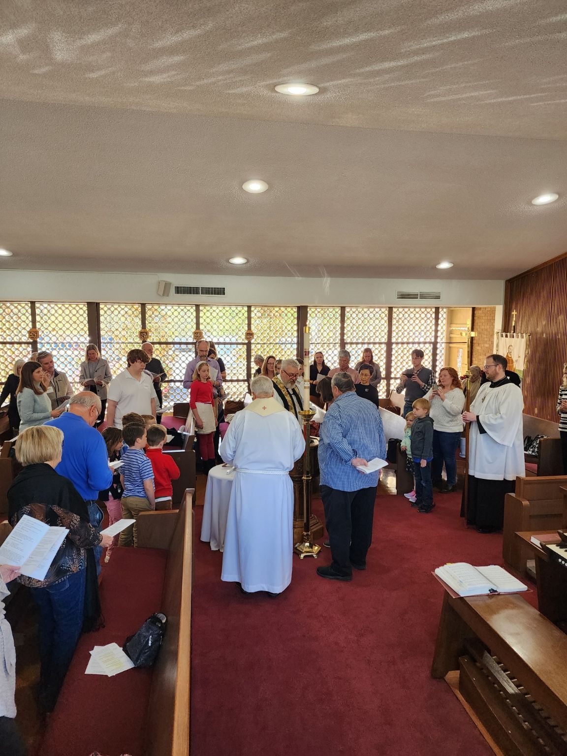 People gathered for a religious ceremony.  A priest in white robe stands at a table. Others hold books, in a church.