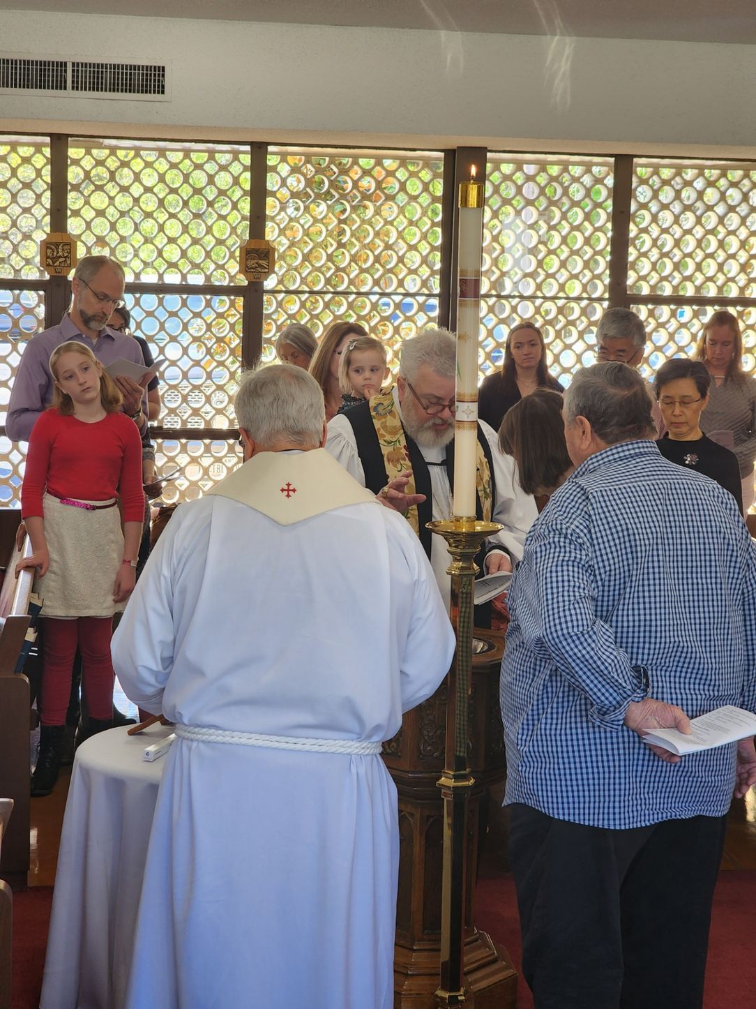 People attending a baptism at a church. A priest pours water. Sunlight streams through a window.