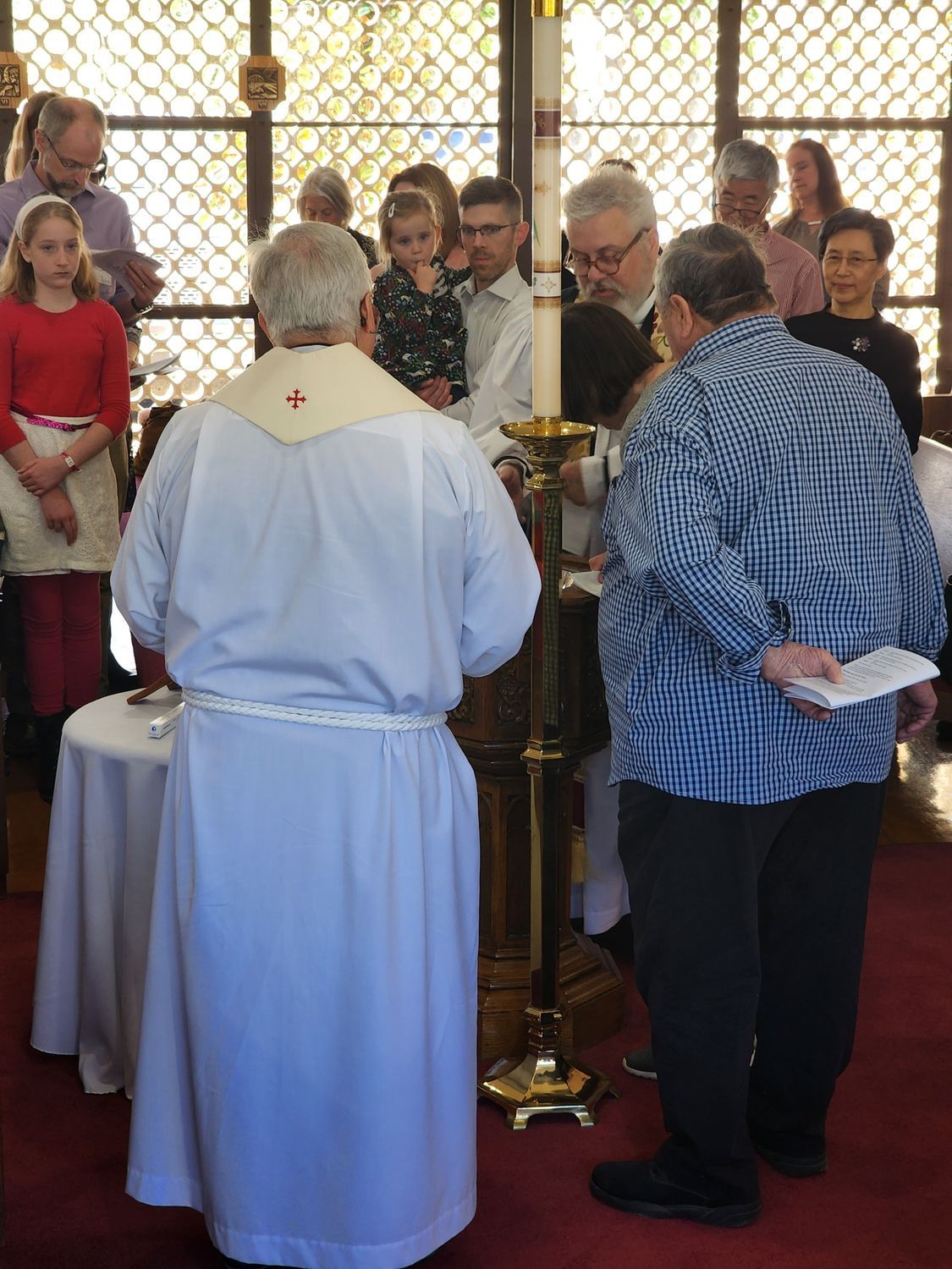 Clergyman lighting a candle during a baptism ceremony in a church, with people watching.