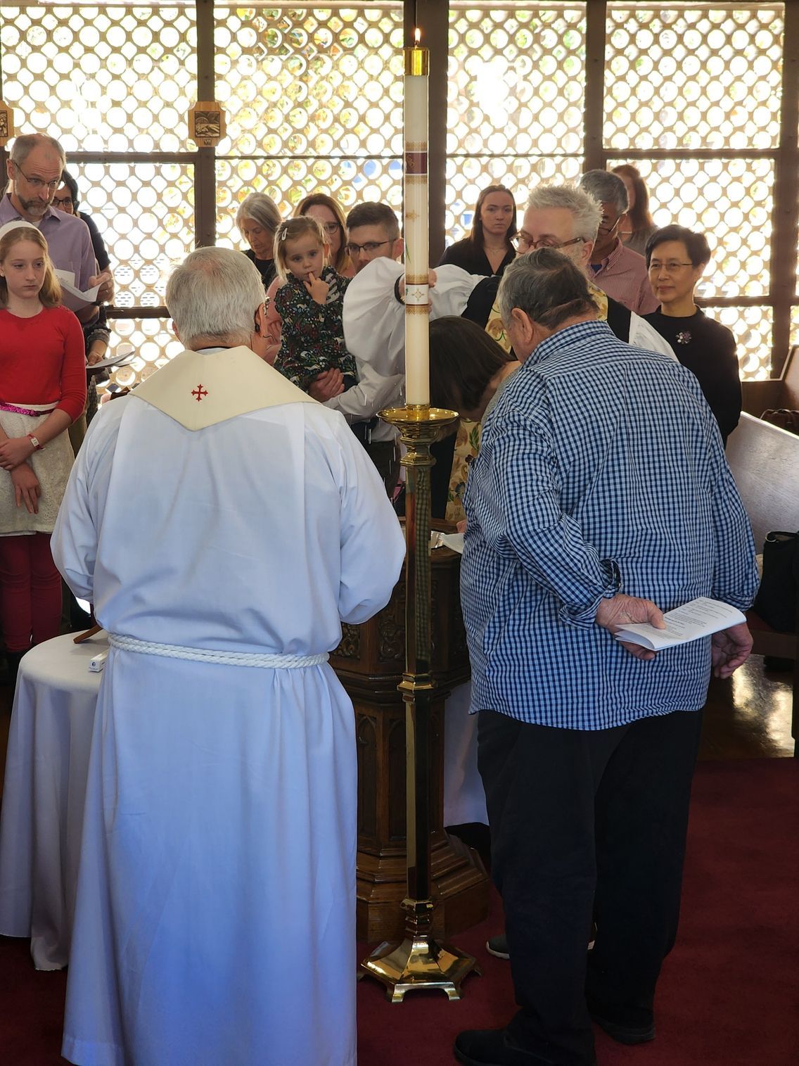 Priest in white robe performs religious ceremony; people gather around a candle in a sunlit church.
