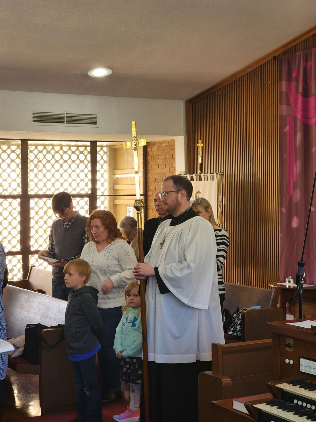 Priest in vestments stands in church, holding a staff, with parishioners nearby.
