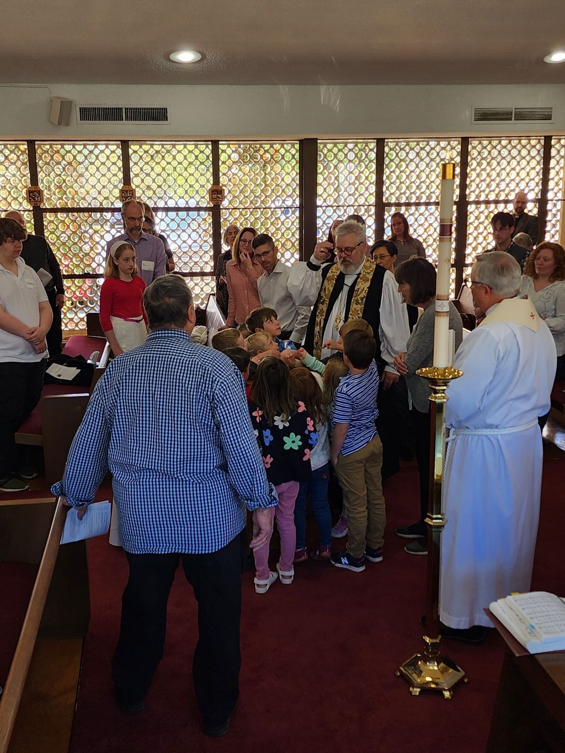 Children gathered with clergy near a candle in a church. People watch in the background.