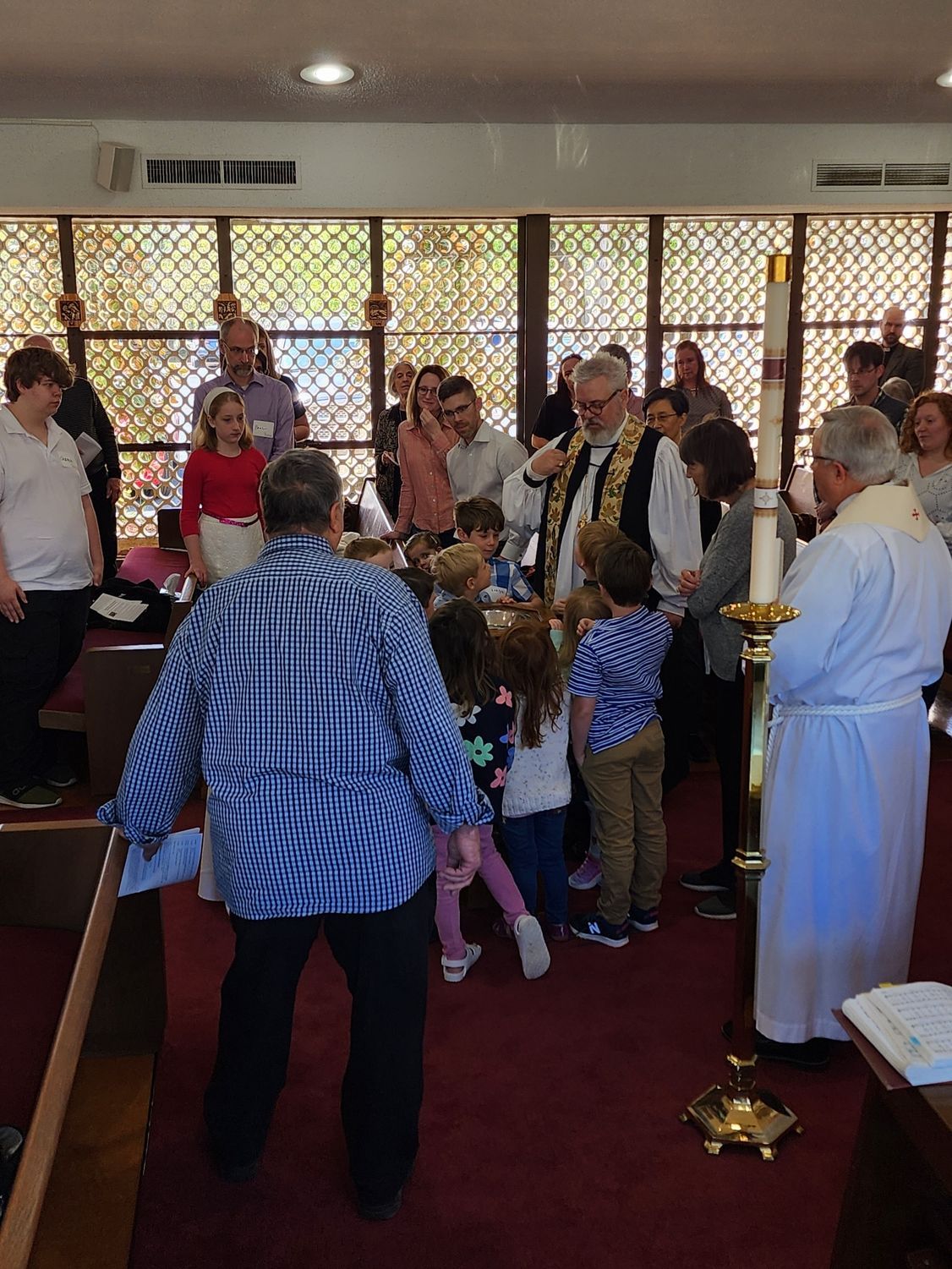 Children and adults in a church, gathered around a priest during a religious ceremony. Sunlight streams through stained glass.