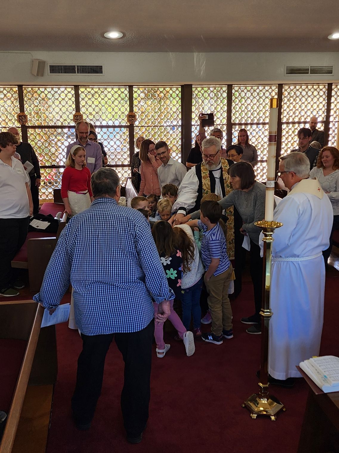 Children at the altar with clergy, possibly a religious ceremony. People surrounding. Bright windows.