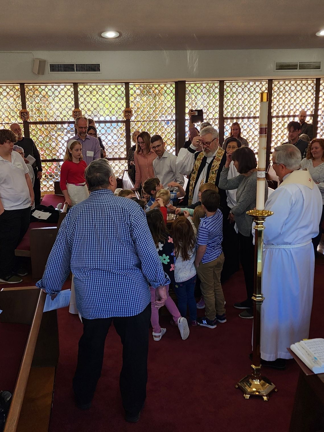 Baptism ceremony in a church: priest pouring water on children, surrounded by people, stained glass windows in background.