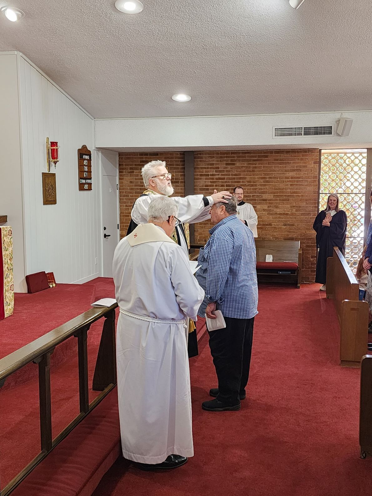 Clergy performing a religious rite, placing a hand on a person's head inside a church with red carpet.