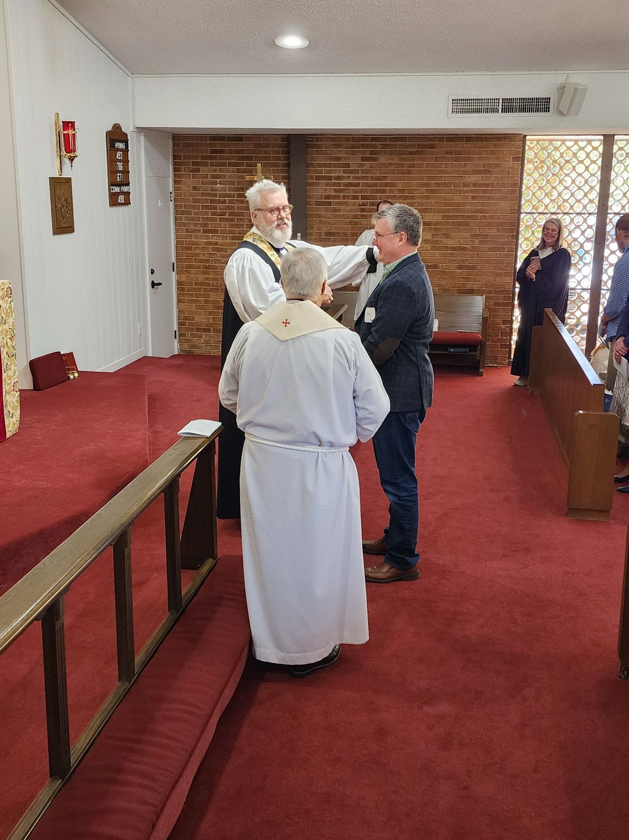 Clergy members at a ceremony, one placing hands on a man's head. Church interior with red carpet and wooden accents.