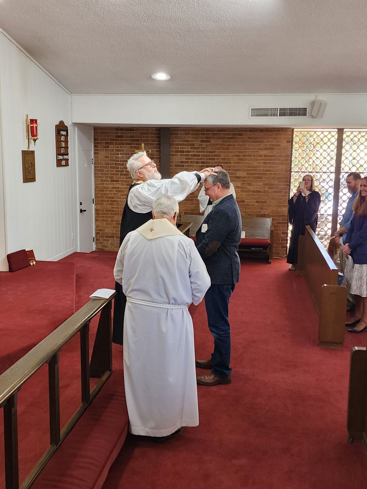 Ordination ceremony in a church: a person is being blessed by a clergyman, with attendees watching.
