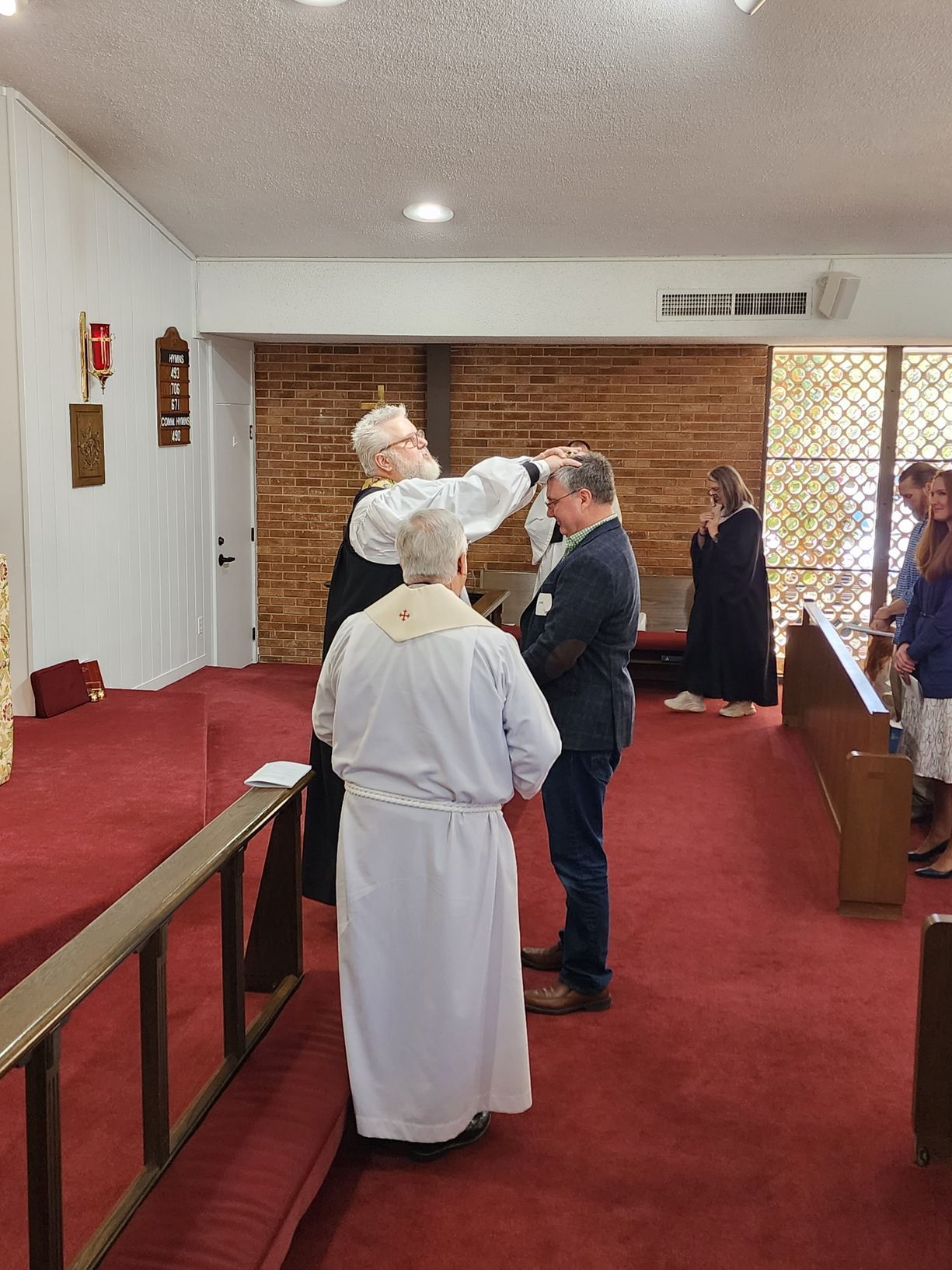 Clergyman anoints a man's forehead in a church, others watch. Red carpet, brick wall, sunlight.