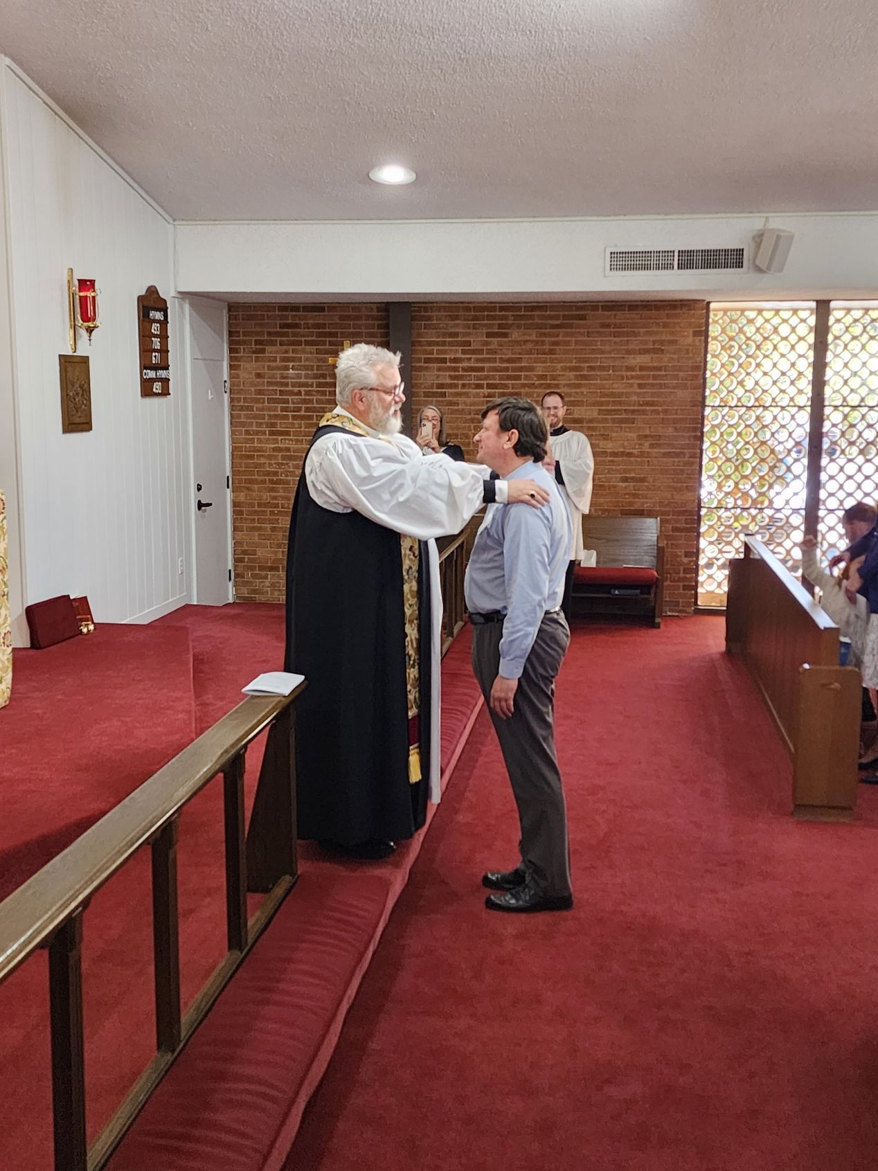 Clergyman in black robes with white collar, laying hands on a man in a church.