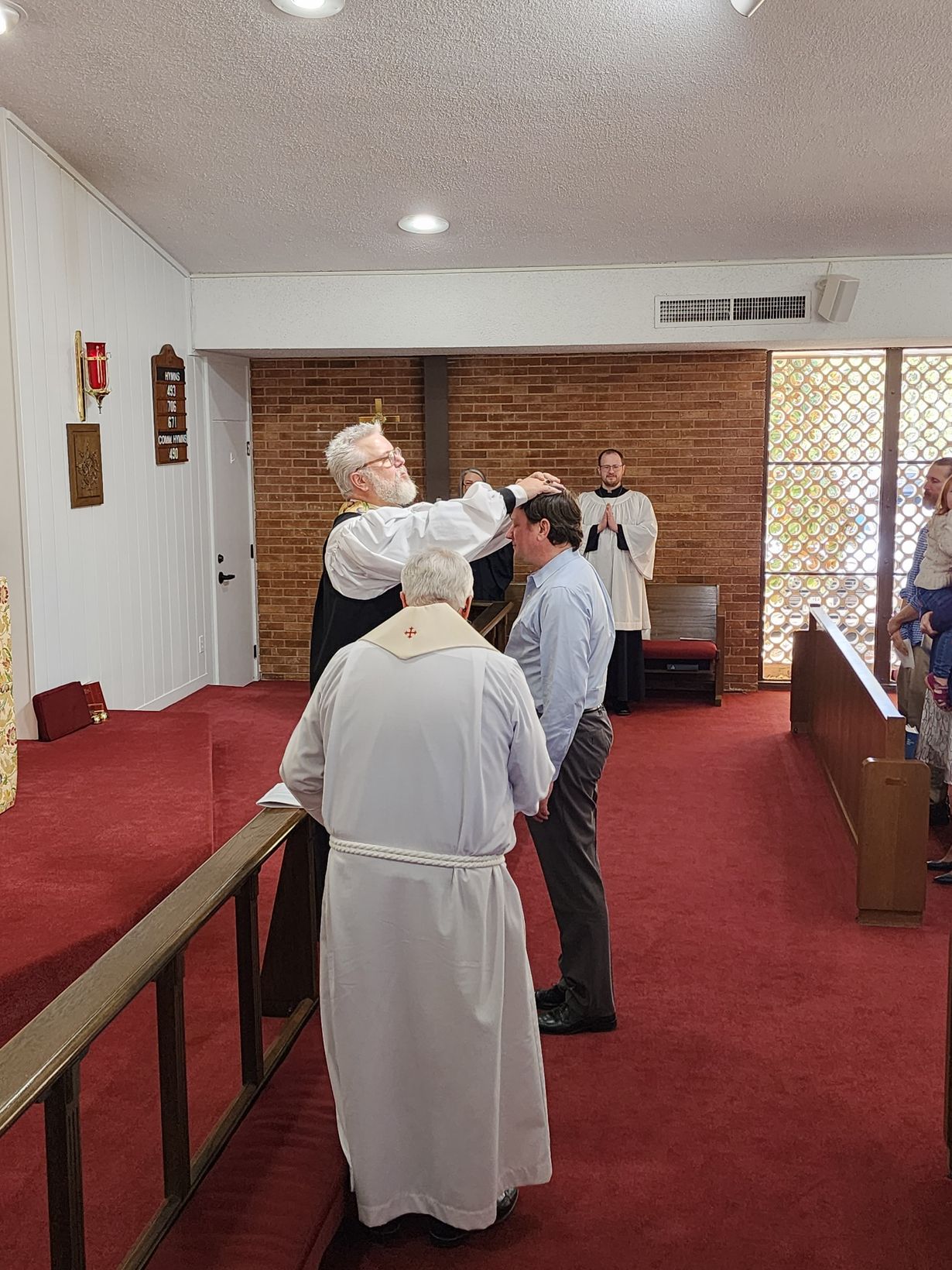 A clergyman anoints a man's forehead in a church with red carpet. Another priest and people observe.