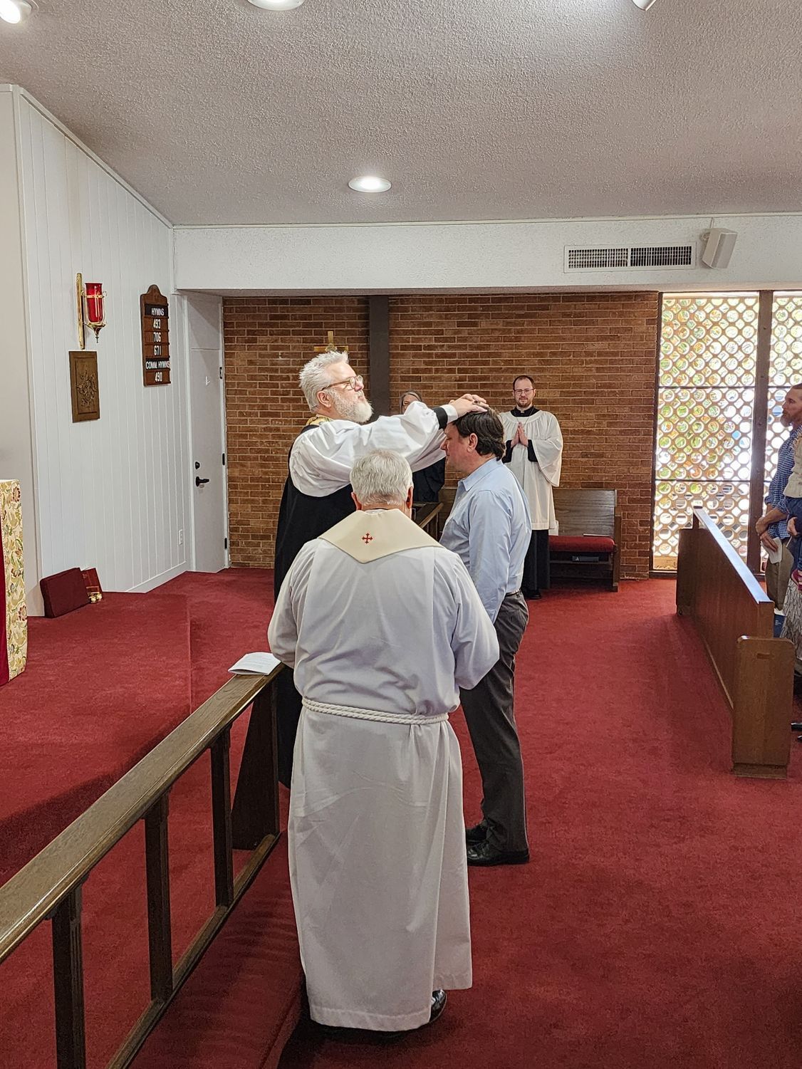 Clergyman anoints a man's forehead during a church service. Others watch, red carpet, light interior.