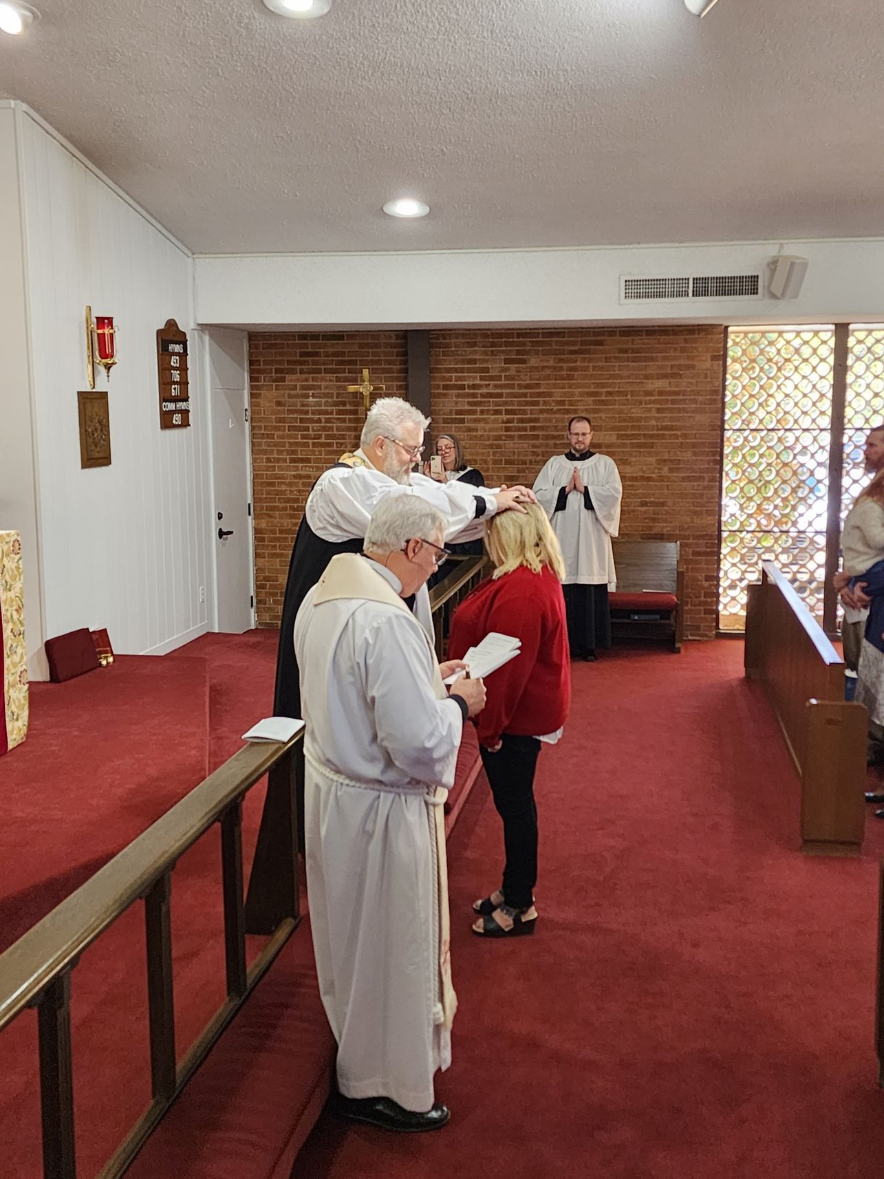 A person being blessed by a cleric in a church. Cleric's hand on the person's head. Others watch. Red carpet.