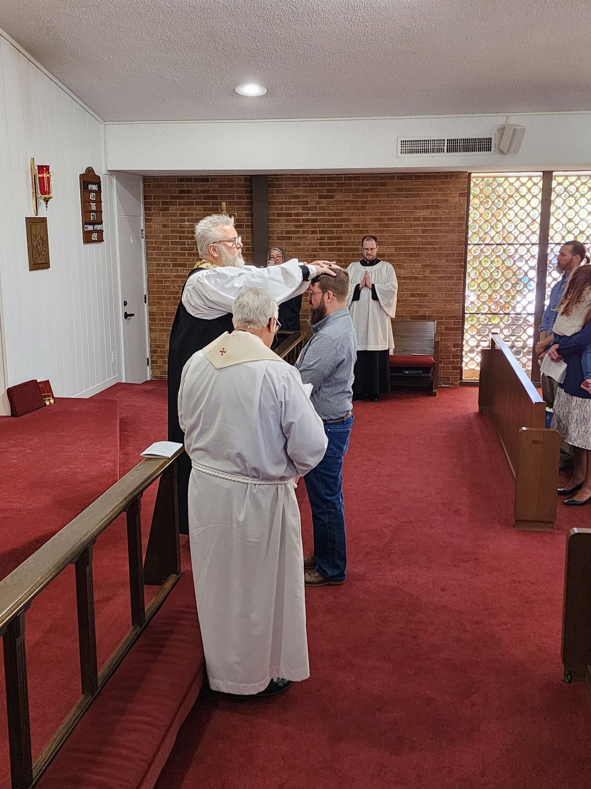 Man being blessed by a clergyman, in a church. Other people watch on red carpet.