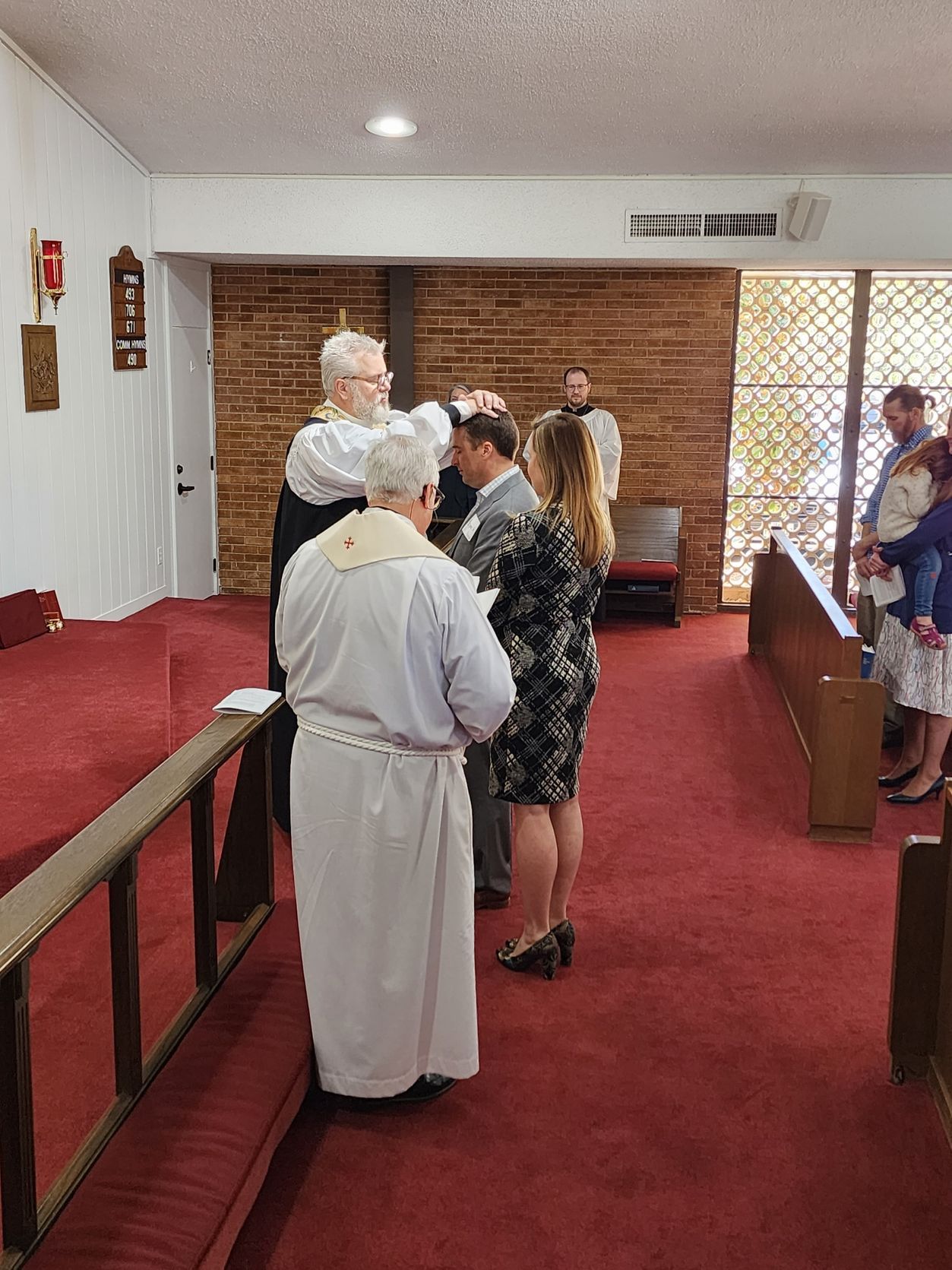 People in church, a ceremony with a person's head touched. Red carpet, light walls, stained glass windows.