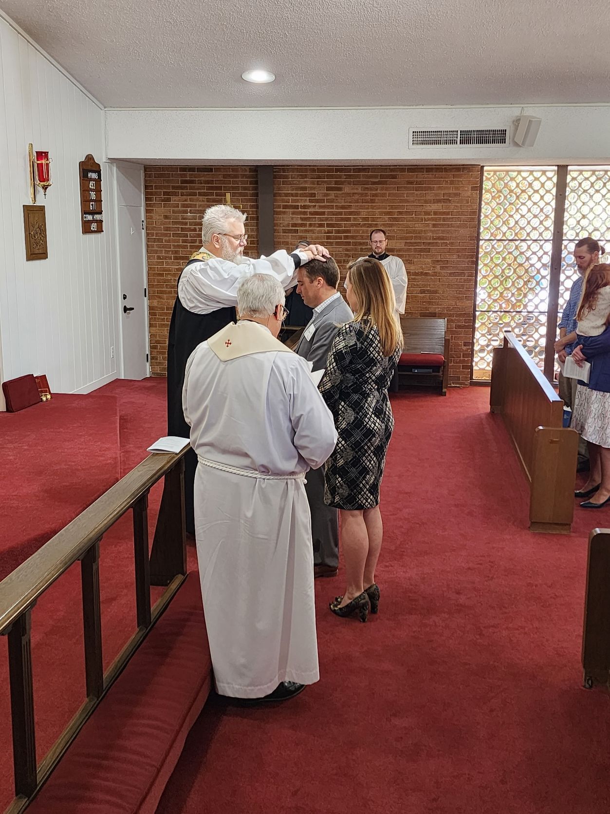 Clergy lay hands on a man during a church ceremony. A woman stands beside him, with others watching.