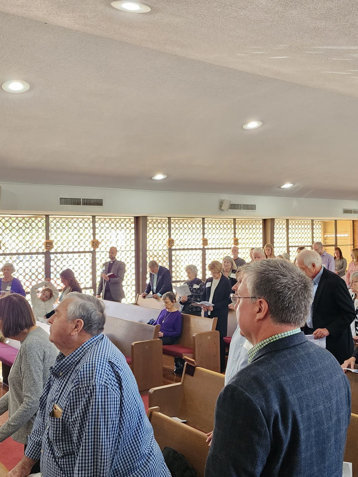People standing in a church service, facing forward. Natural light enters through side windows.