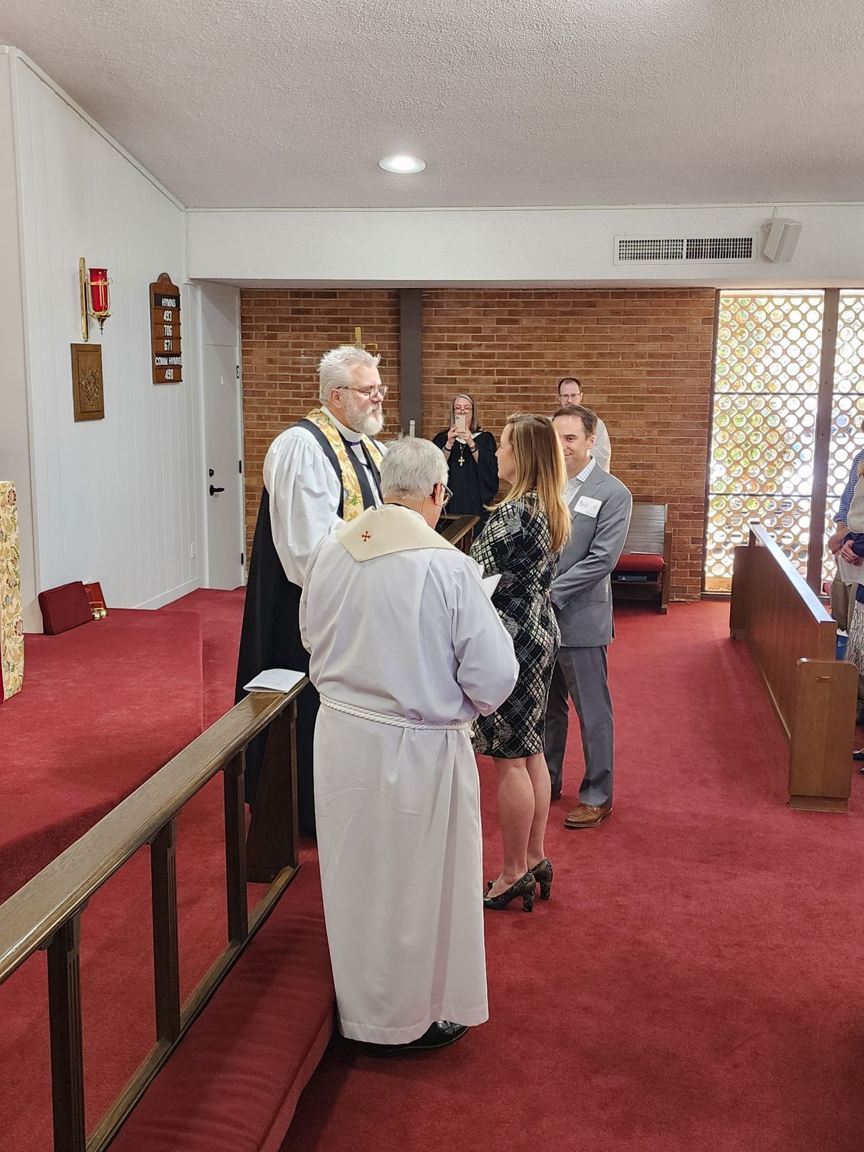 Wedding ceremony in a church. A priest and couple stand facing each other, officiant in vestments, red carpet.