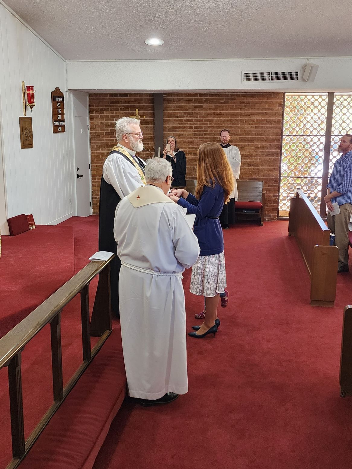 Clergy perform a religious ceremony. A woman in a floral skirt stands before them in a church. Red carpet and wooden pews.