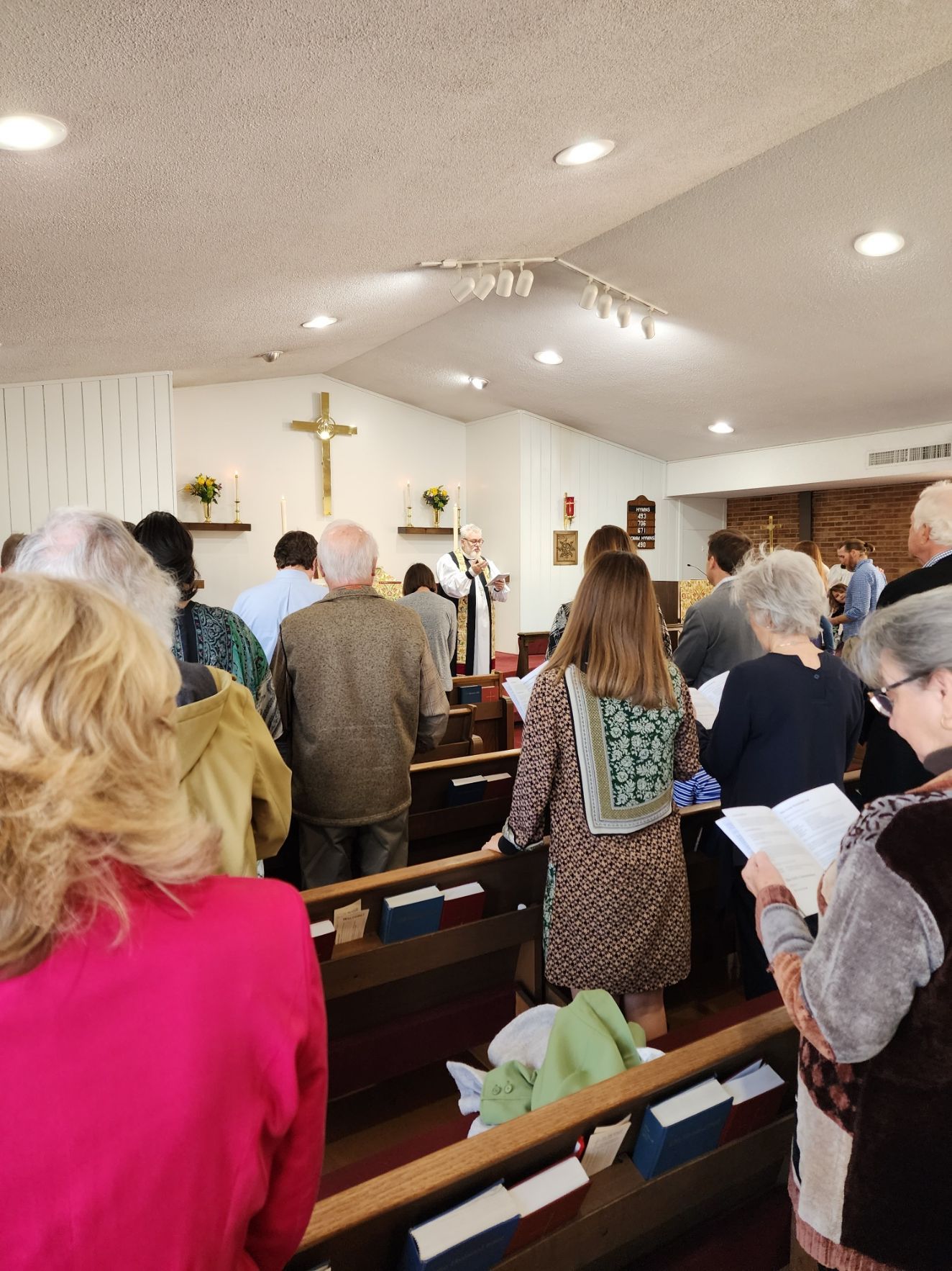 People standing and facing forward in a church, listening to a service. A cross and altar are visible.