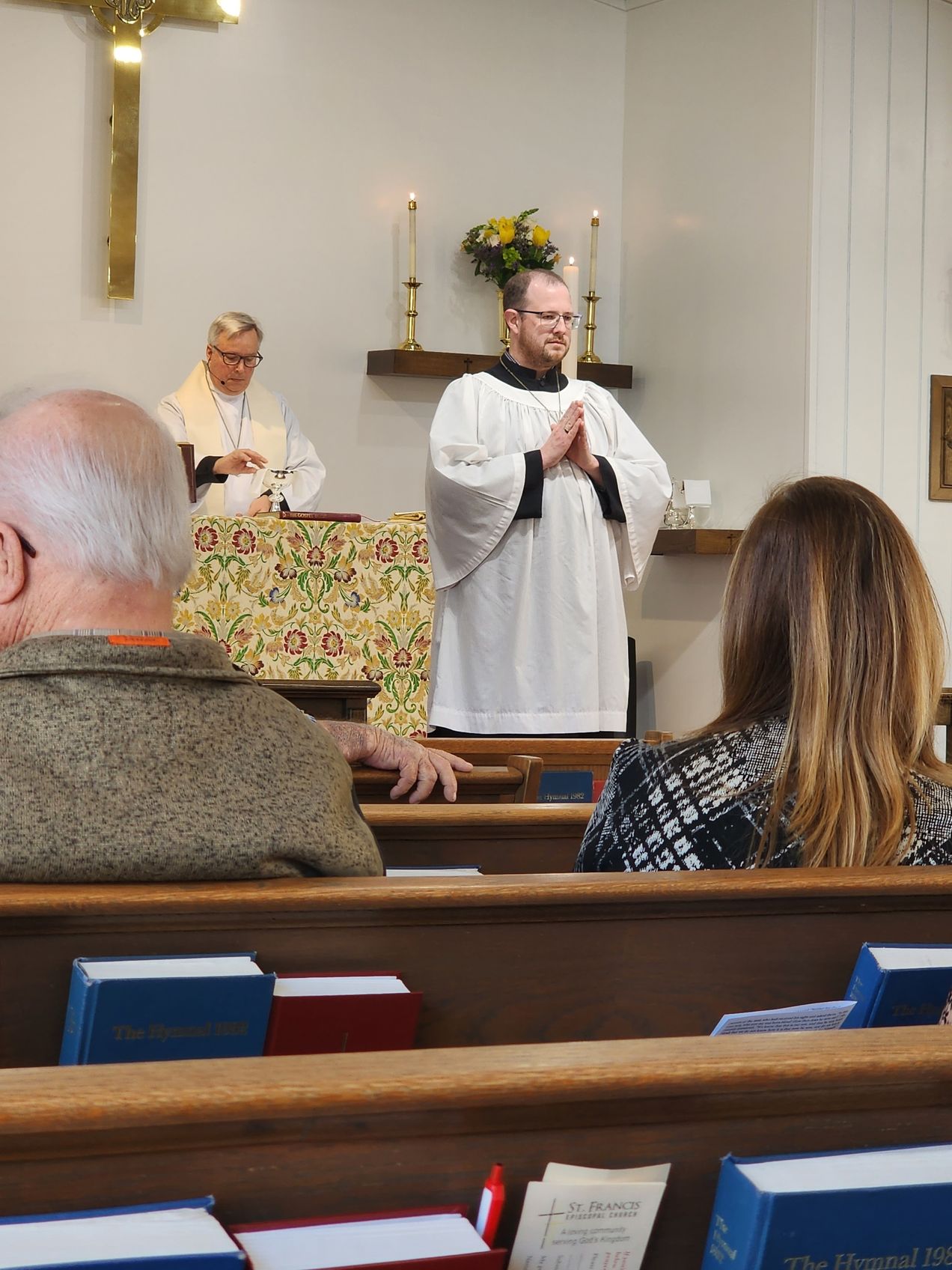 A church service: A man in a white robe leads prayer, others seated in pews, another priest at the altar.