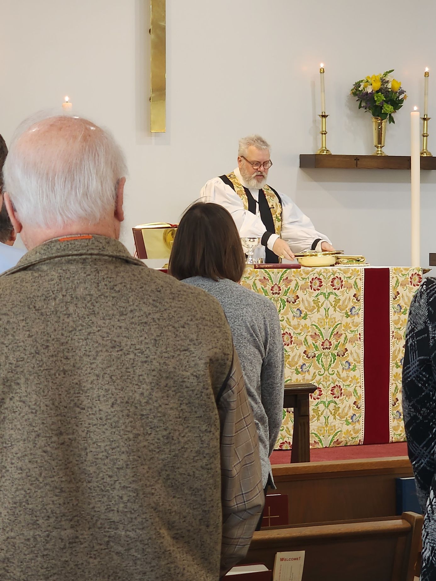 People in church observe a service. A priest in vestments stands at the altar.