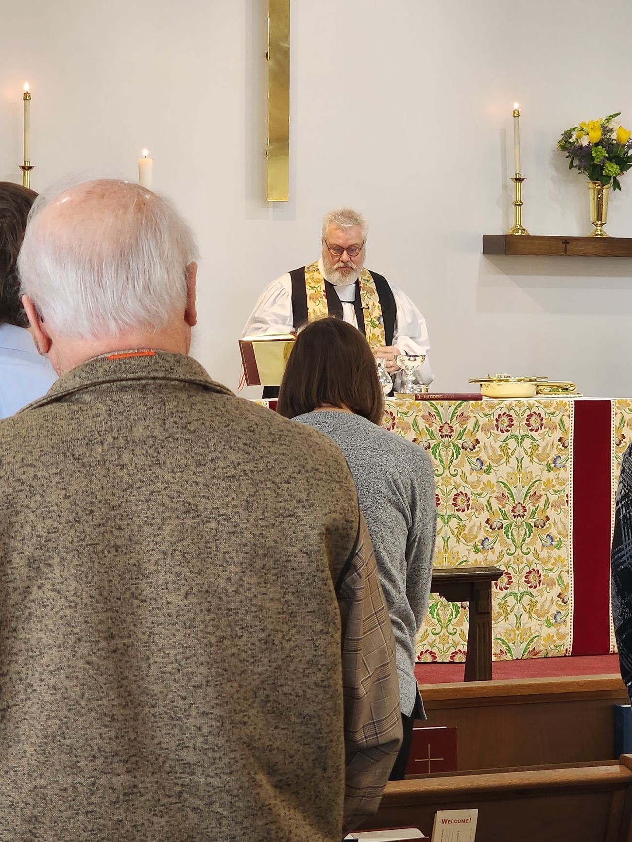 People in church, facing a robed officiant at the altar. Two candles and floral decoration.