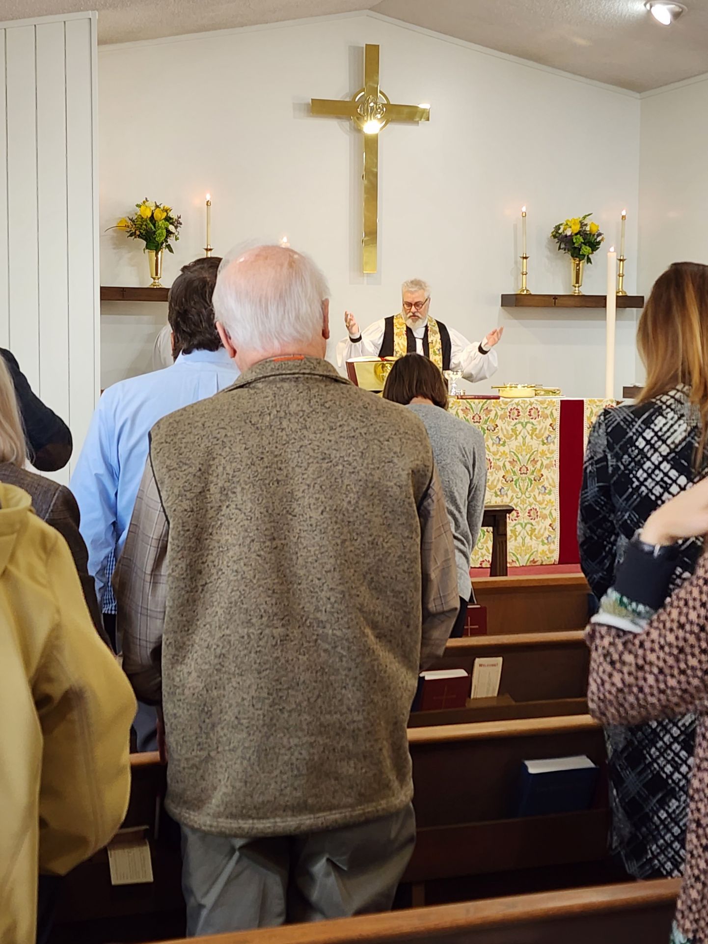 People in church facing altar with cross; a clergyman is at the altar, speaking with arms raised.