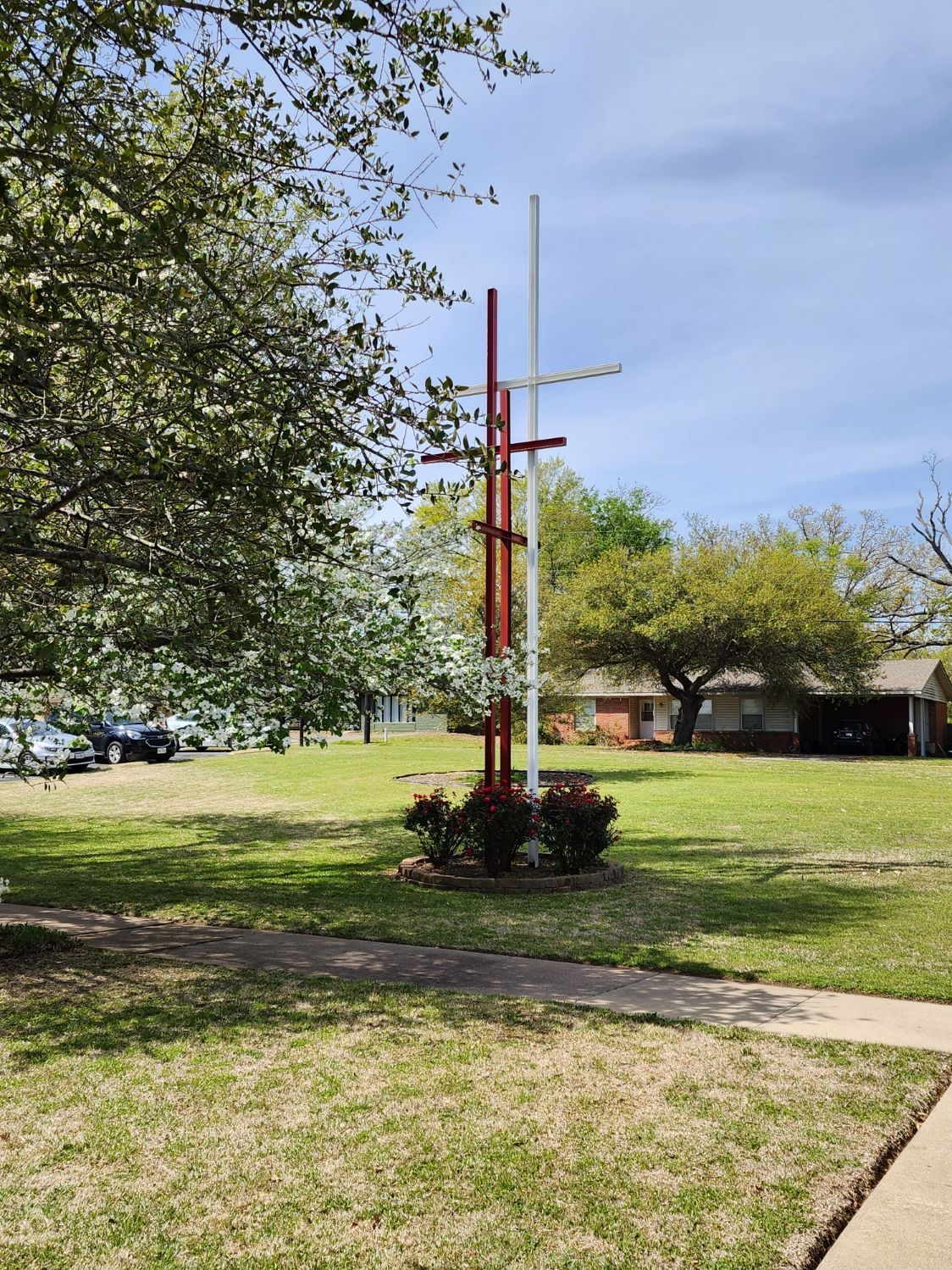 Tall, red and white cross sculpture in a grassy yard, trees and houses in the background.