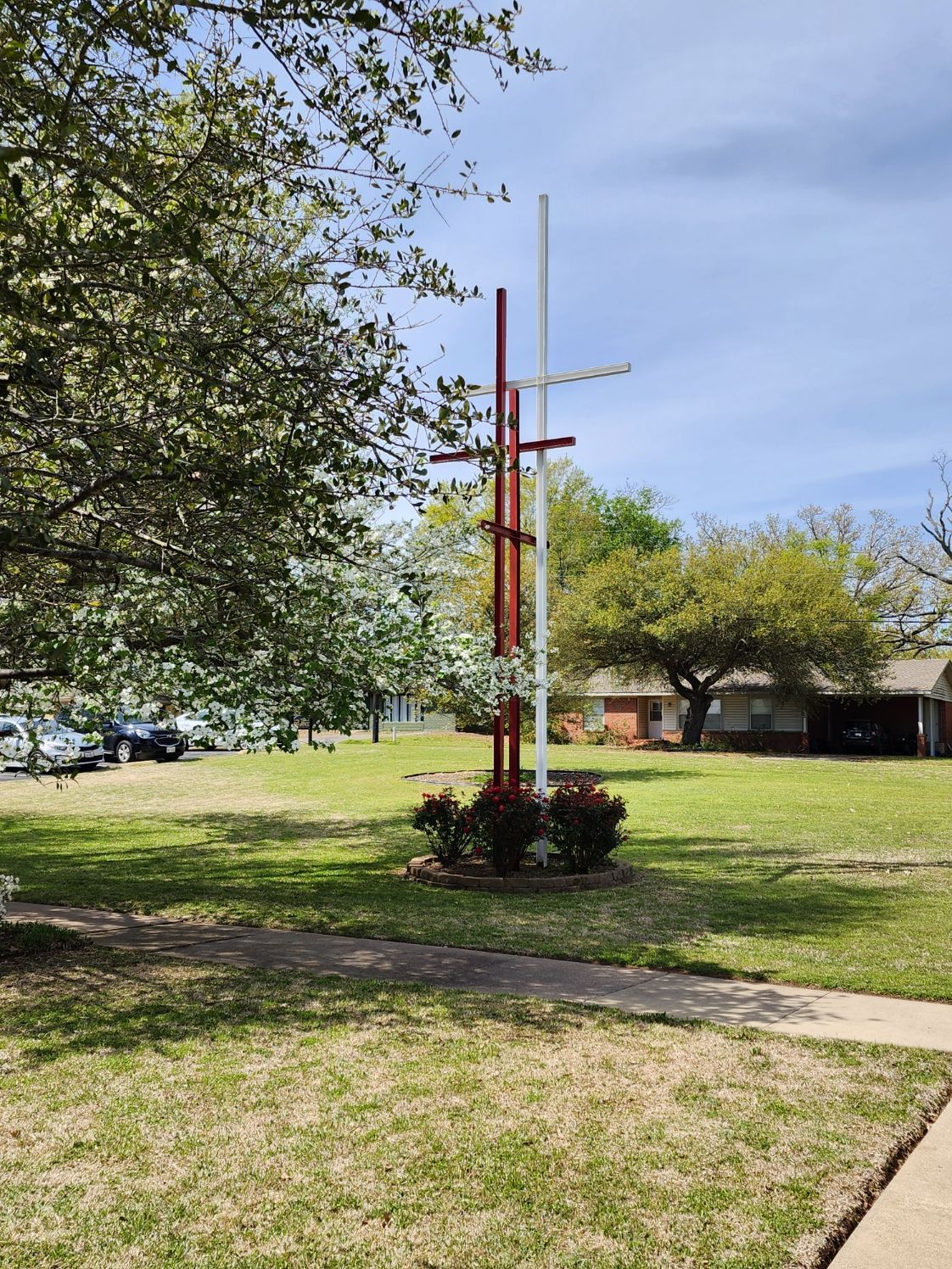 Tall, artistic, red and white metal sculpture in a grassy park, next to trees and a building.