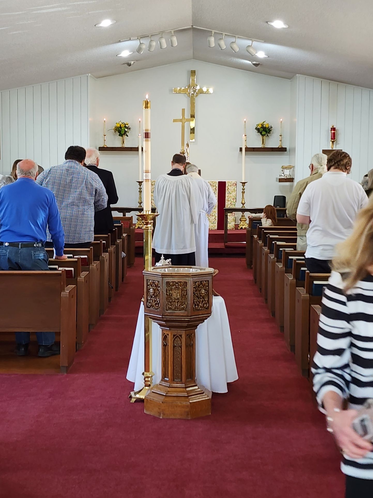 Church service: Priest at altar, people in pews, wooden baptismal font, cross, candles.