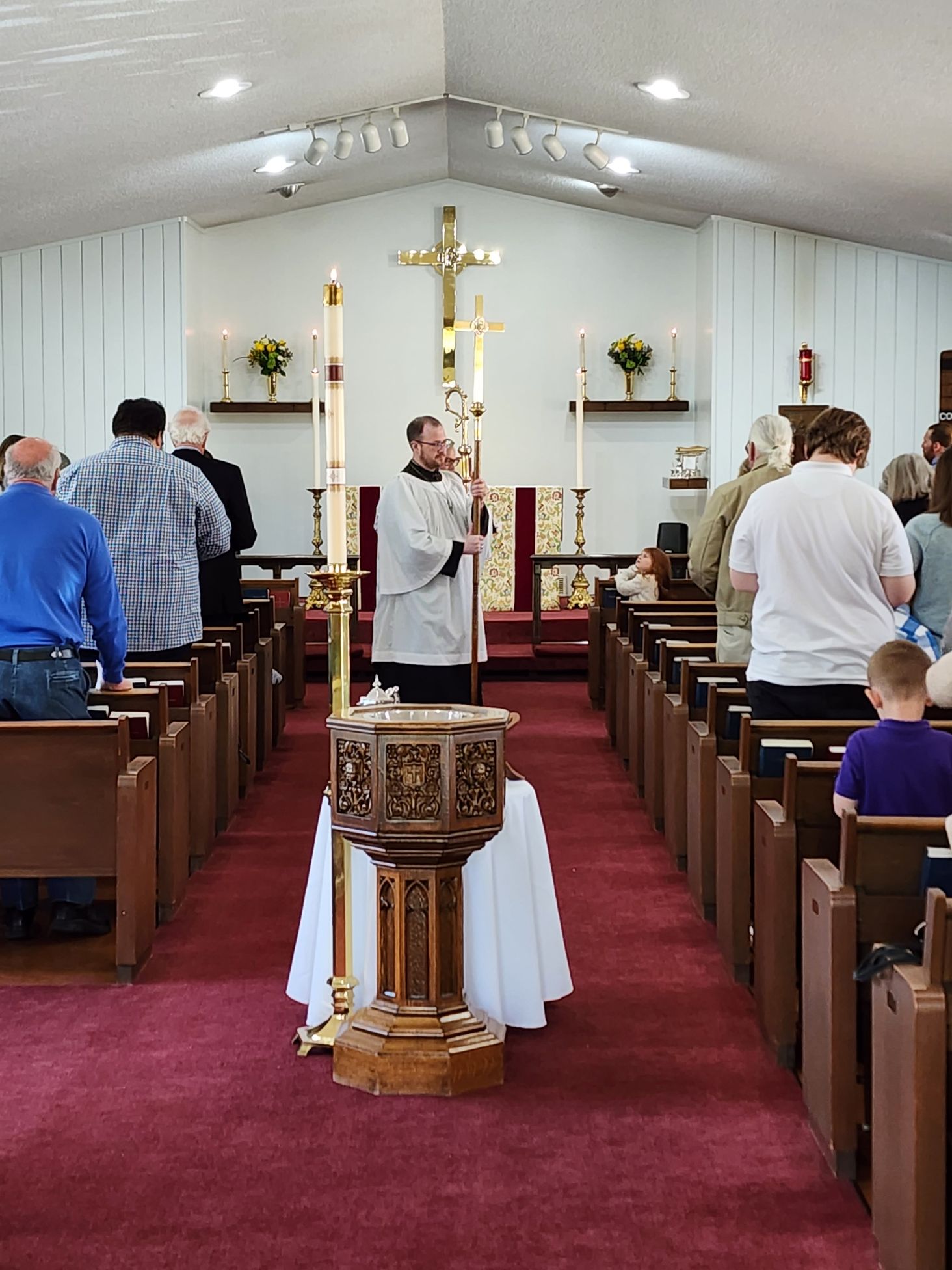 Church service in progress: priest holding a cross, people standing, pews, altar with a crucifix, candles lit.