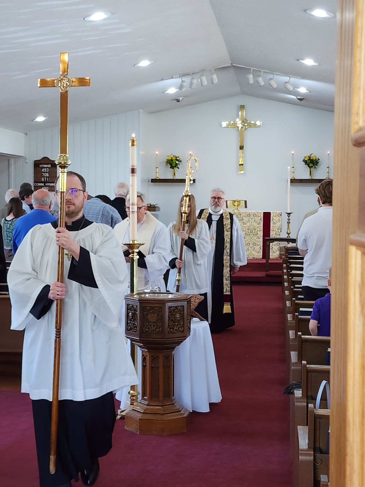 Clergy process down church aisle. People carry cross, candles. Red carpet, wooden altar.