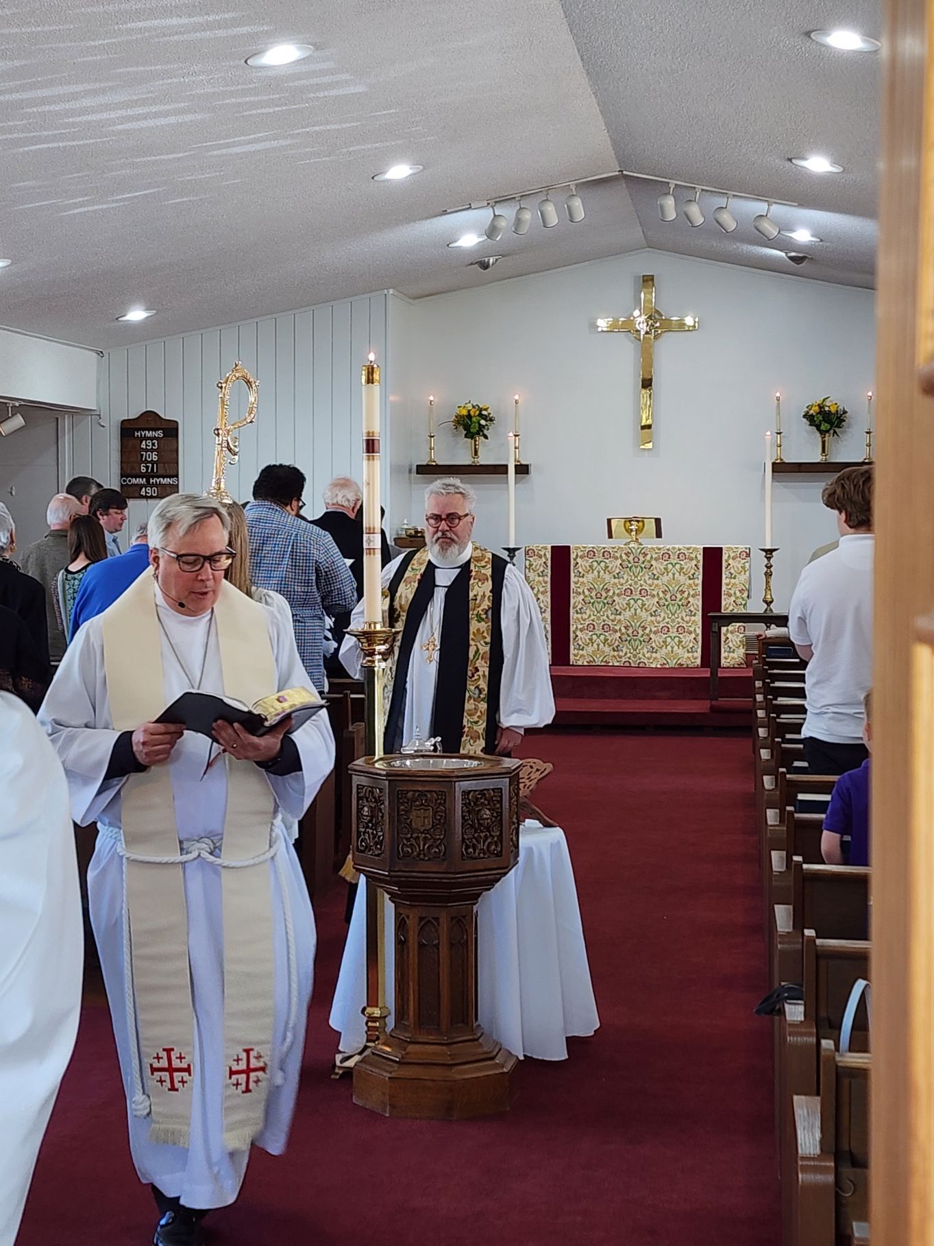 Clergy in robes walk inside a church. A baptismal font, altar, and cross are visible.