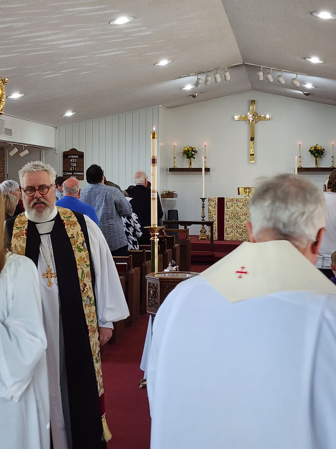 Inside a church, clergy in vestments, some with crosses, stand with other people near an altar, gold crucifix visible.
