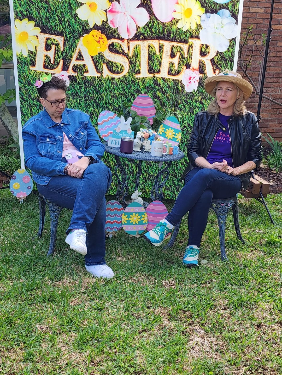 Two women sitting outdoors by Easter decorations. One wears a denim jacket and glasses, the other a jacket and straw hat.