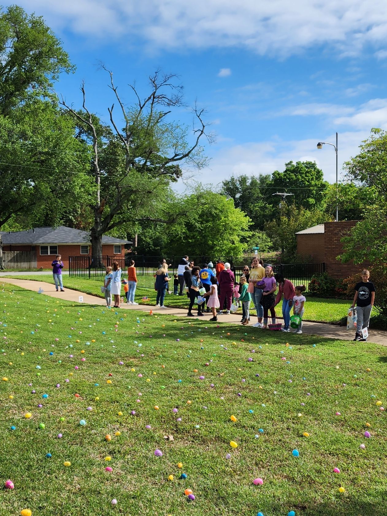 Children and adults gathering outdoors for an Easter egg hunt on a grassy field with colorful eggs scattered.