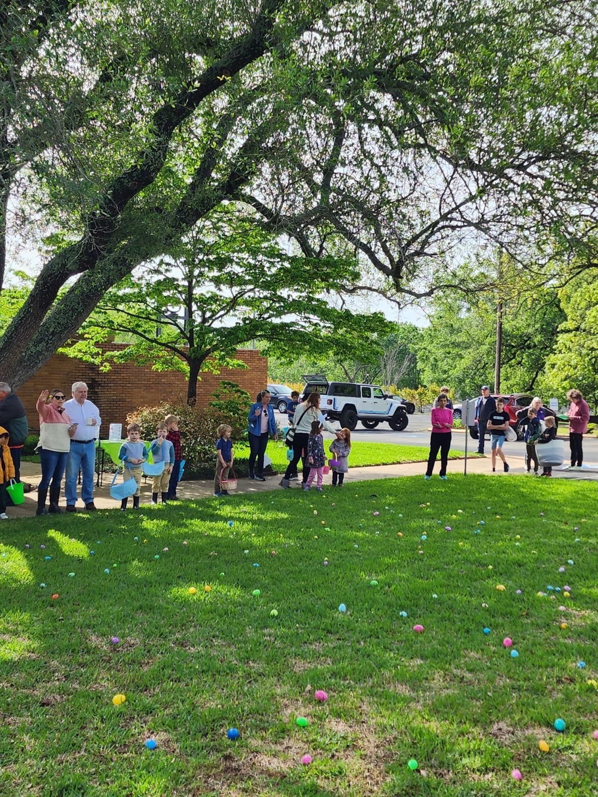 People gather on a grassy field, likely for an Easter egg hunt. Colored eggs dot the green lawn. A large tree shades the scene.
