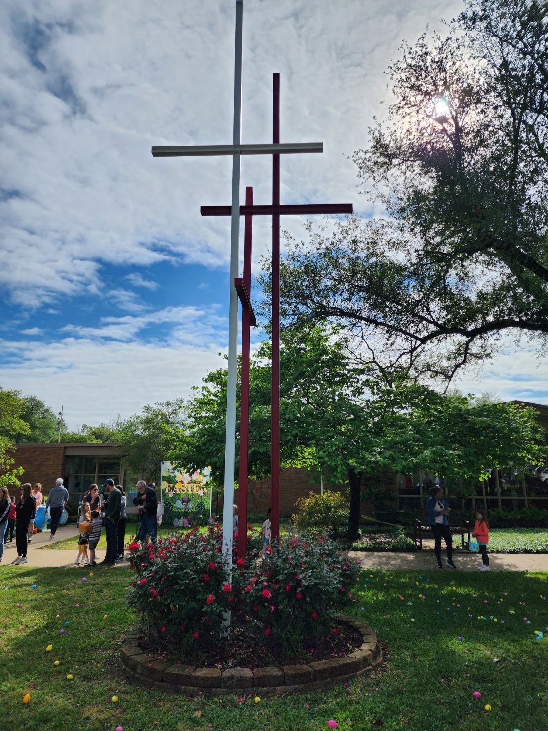 Three crosses stand outside a building; people gather on a sunny day.