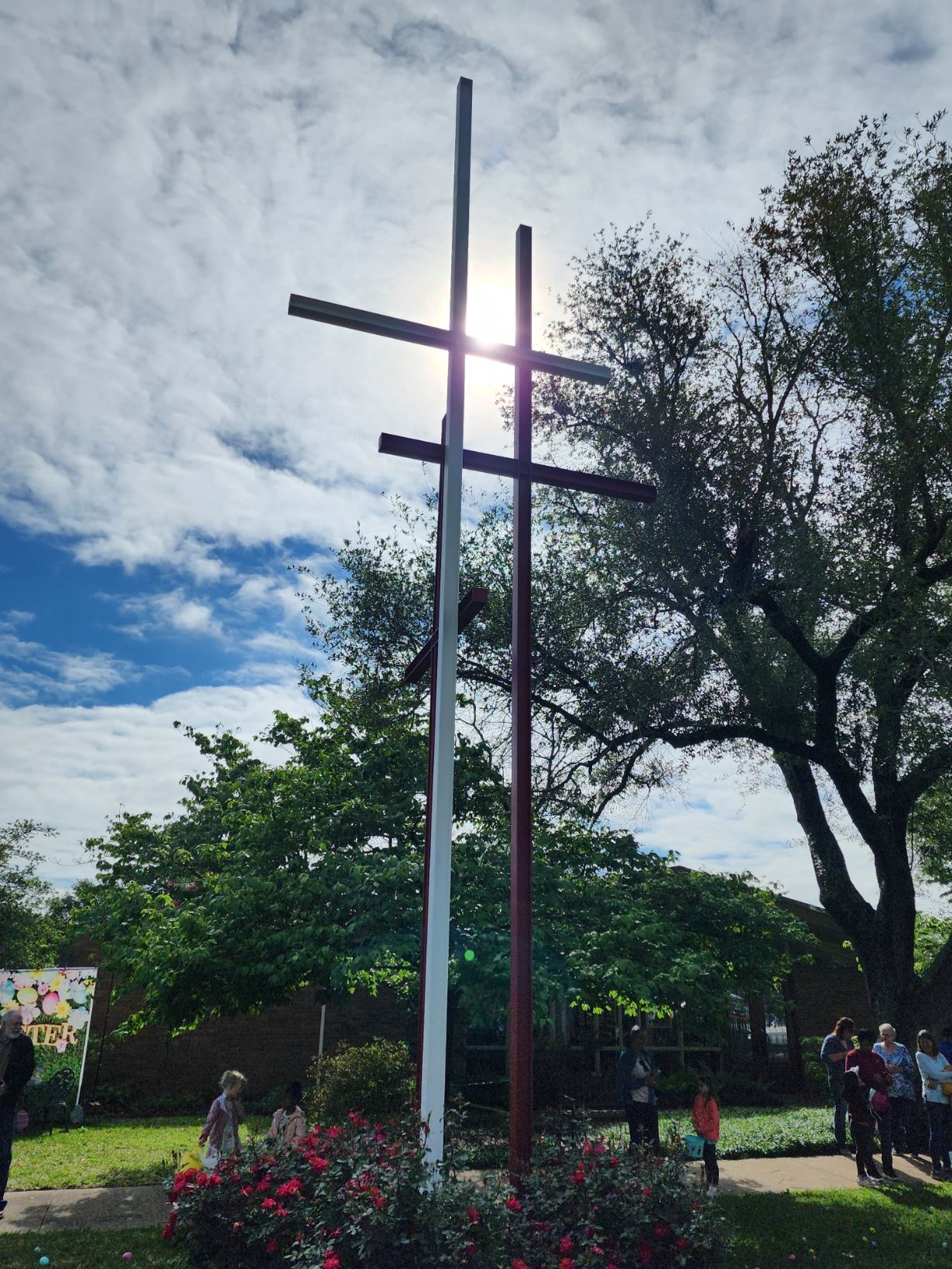 Three crosses stand in front of a sunny sky and lush green trees; people gather below.